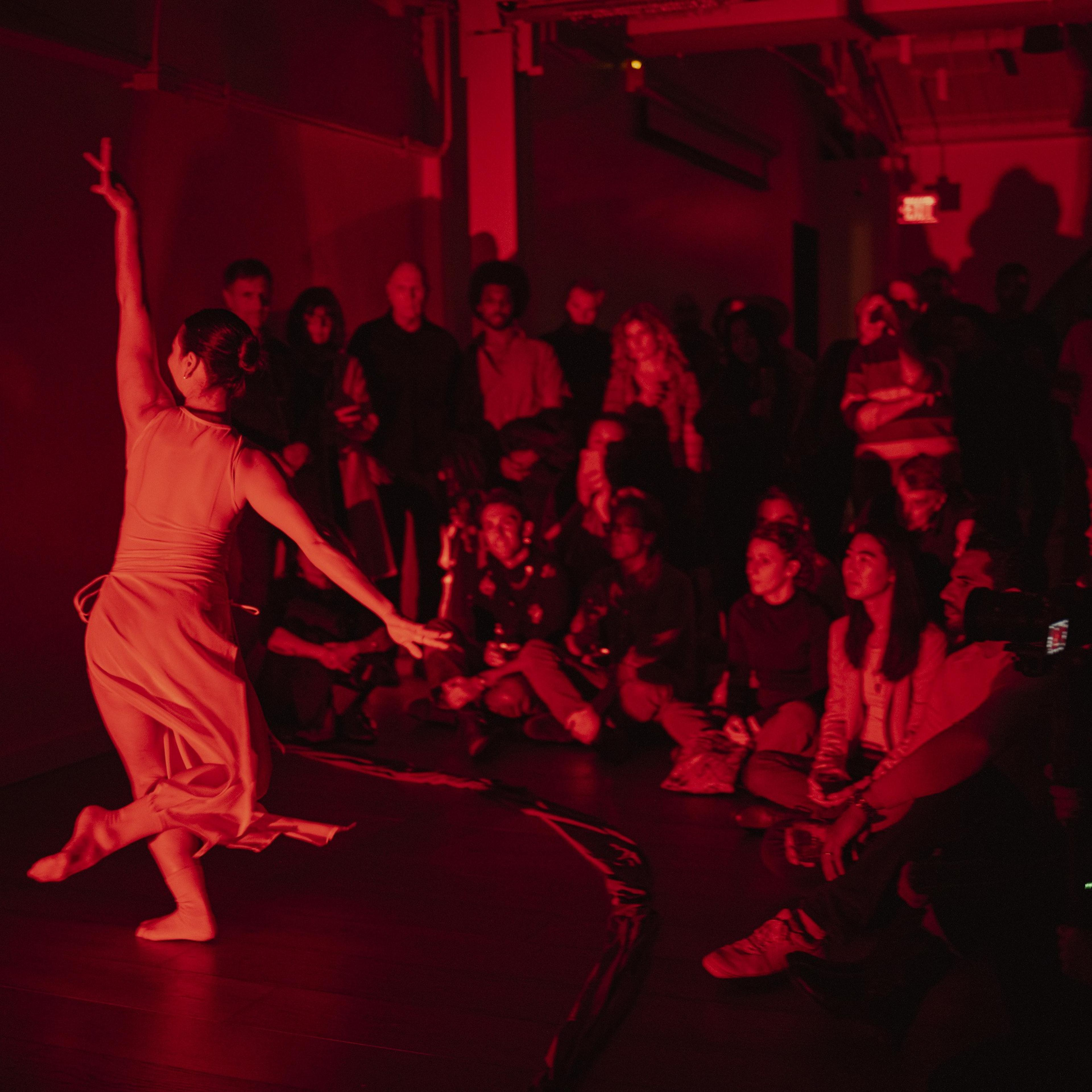 A dancer is performing in a dimly lit room bathed in red light, while a crowd of people attentively watches, some seated on the floor and others standing.