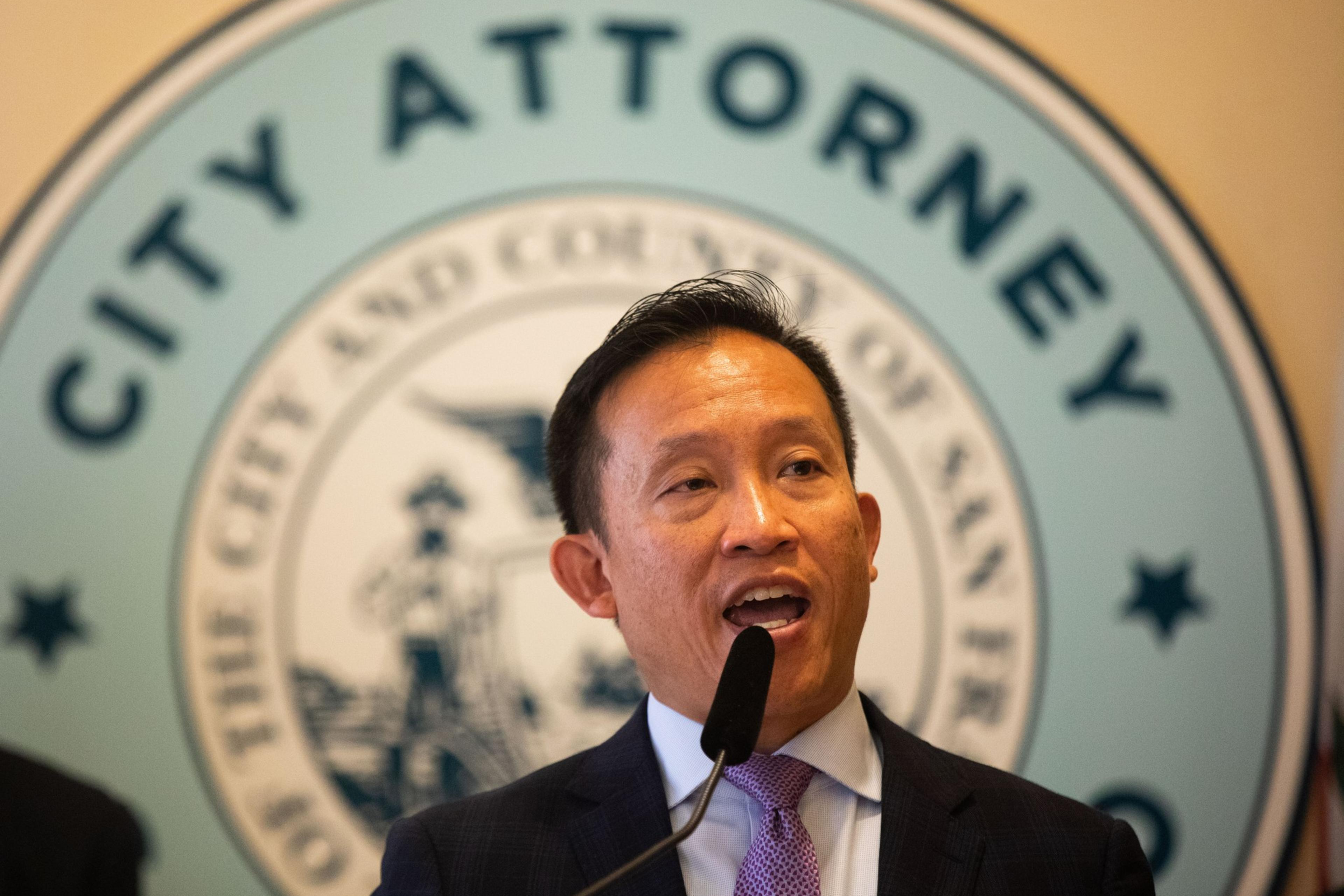 A man in a suit and tie speaks at a microphone, in front of a sign that reads "City Attorney" and features the seal of San Francisco.