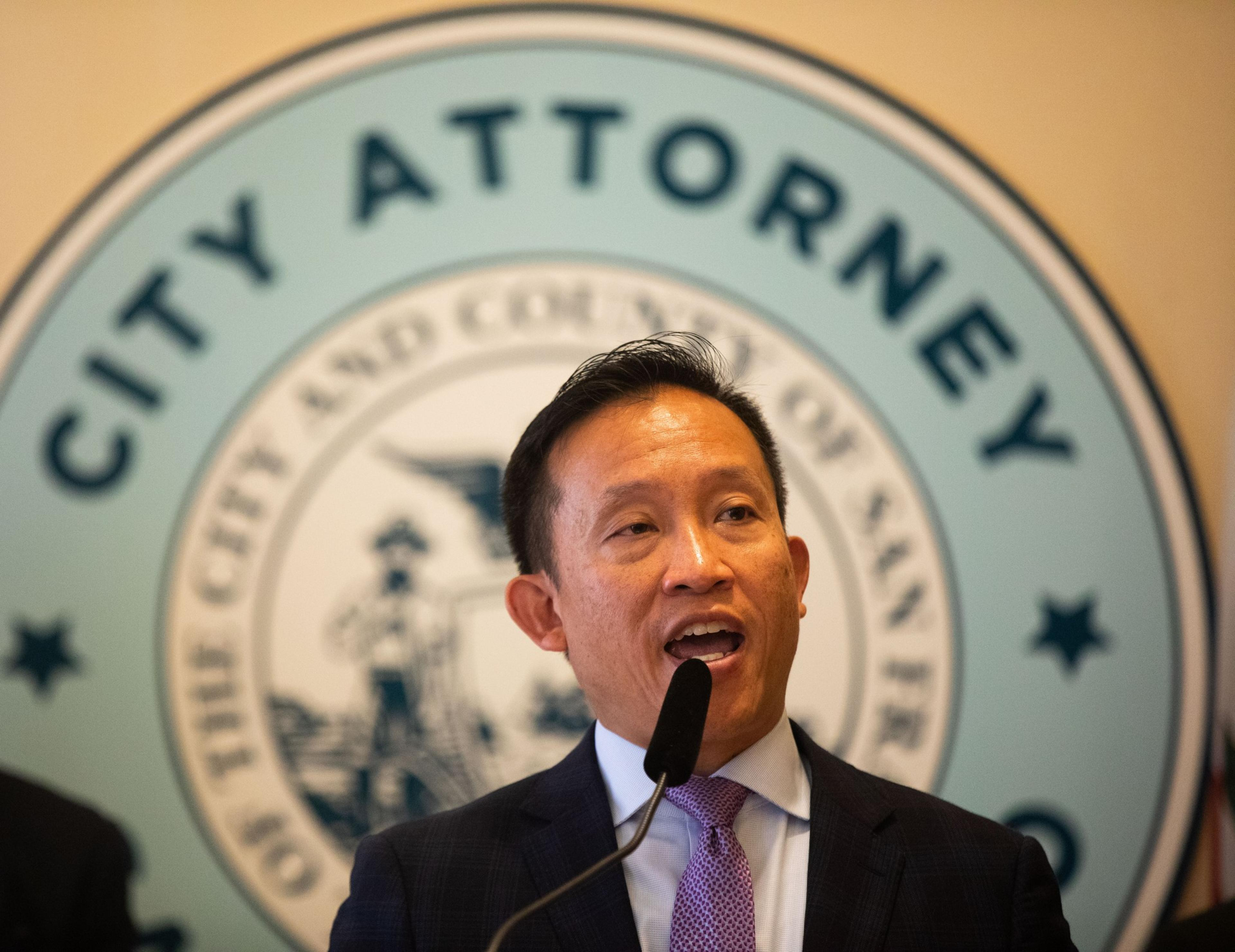 A man in a suit and tie speaks at a microphone, in front of a sign that reads &quot;City Attorney&quot; and features the seal of San Francisco.