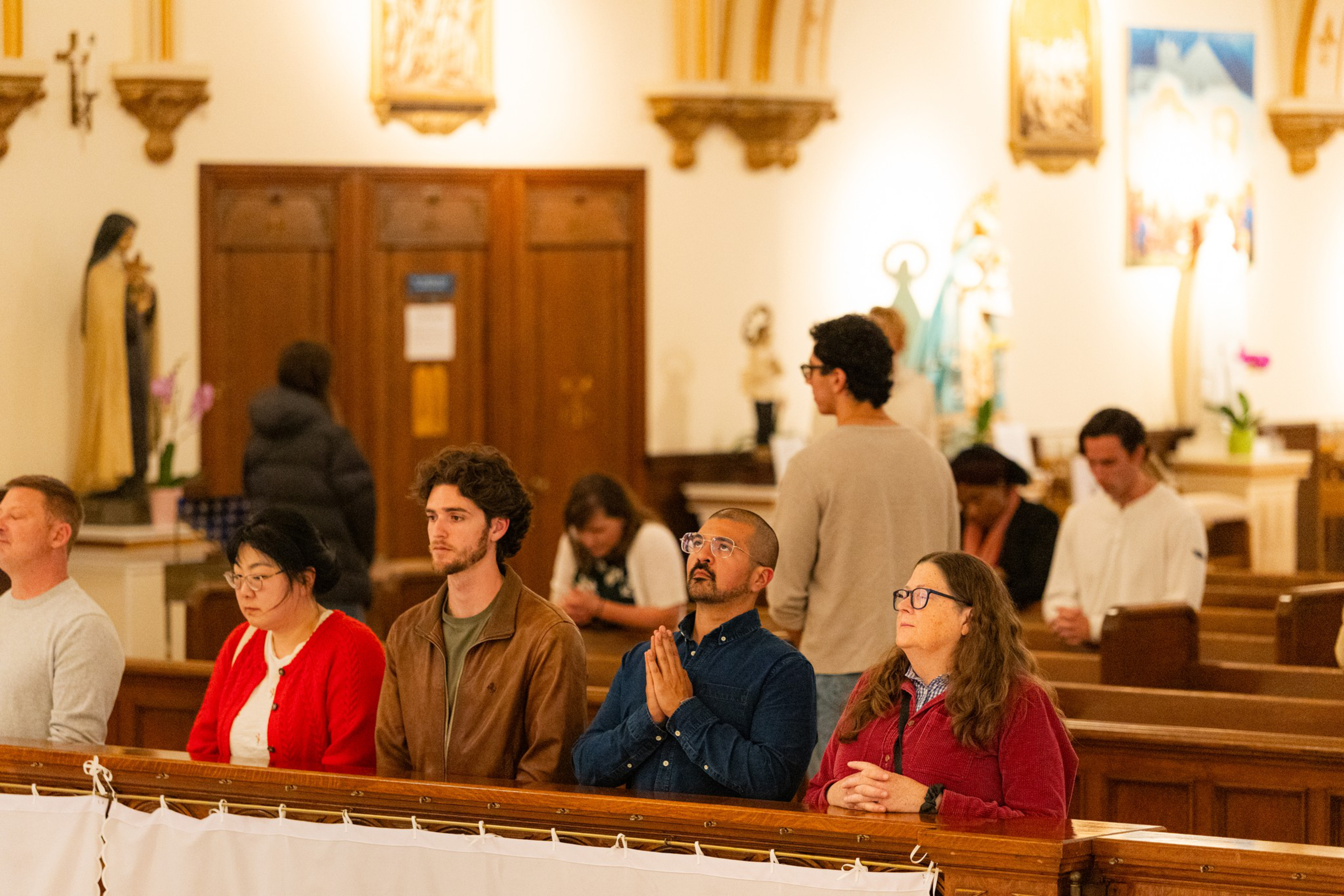 People of diverse backgrounds sit and stand inside a church, some with heads bowed in prayer and others looking forward attentively.