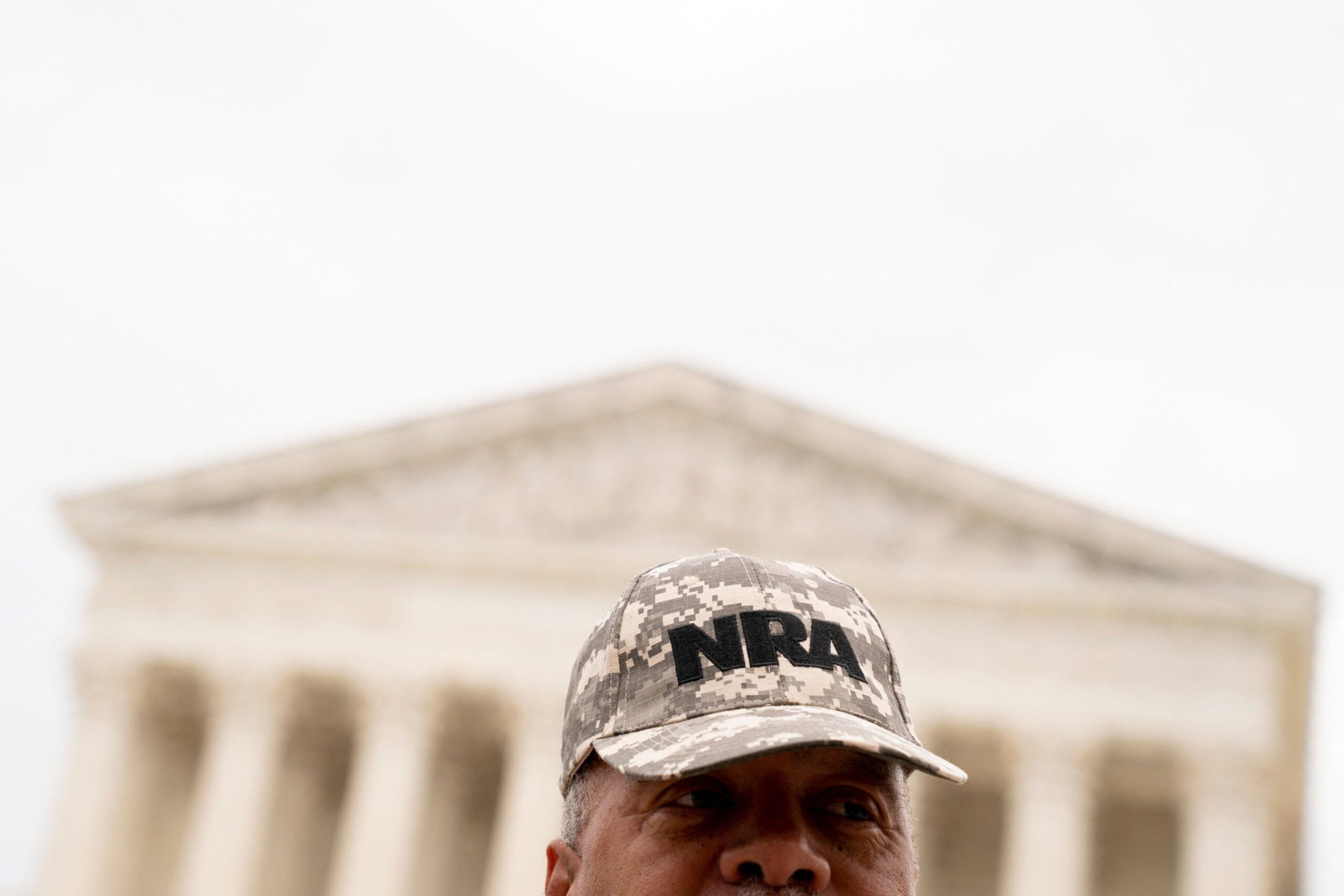 A man wearing a camouflage cap with "NRA" embroidered on it stands in front of a large neoclassical building with tall columns.