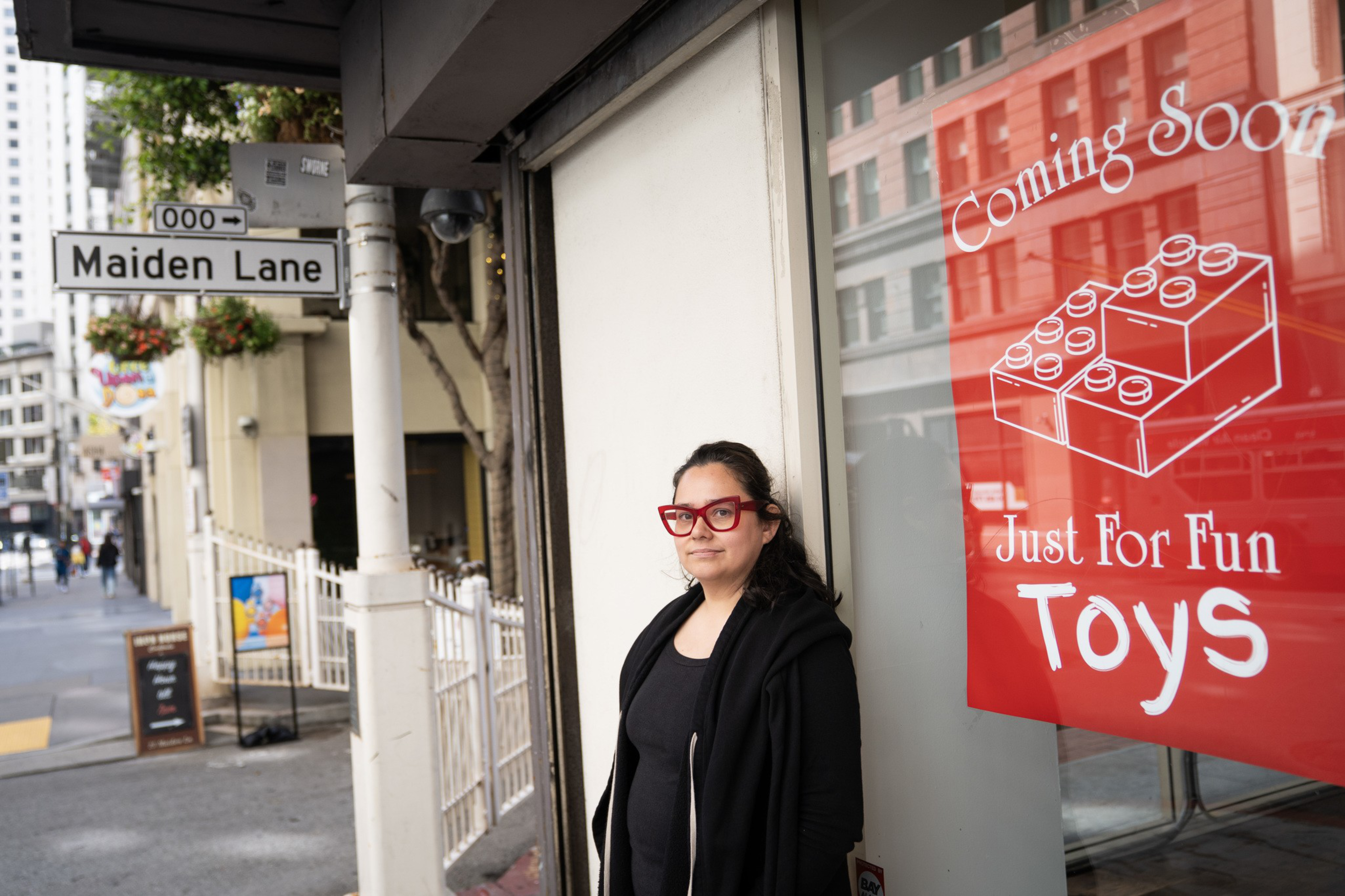 A woman with red glasses and a black outfit stands near a store window with a red sign that reads “Coming Soon Just For Fun Toys” on Maiden Lane street.