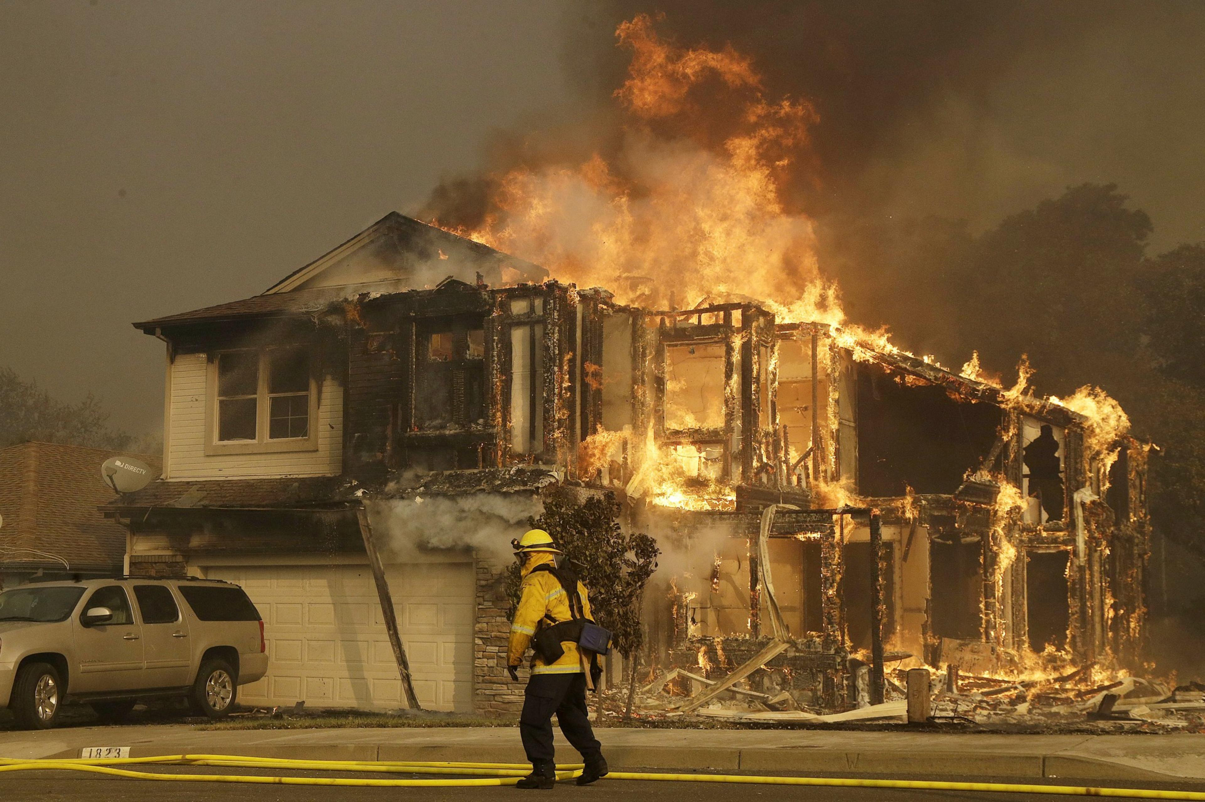 A house is engulfed in flames, severely damaged, with intense fire and smoke. In the foreground, a firefighter in yellow gear walks by, surrounded by yellow hoses.