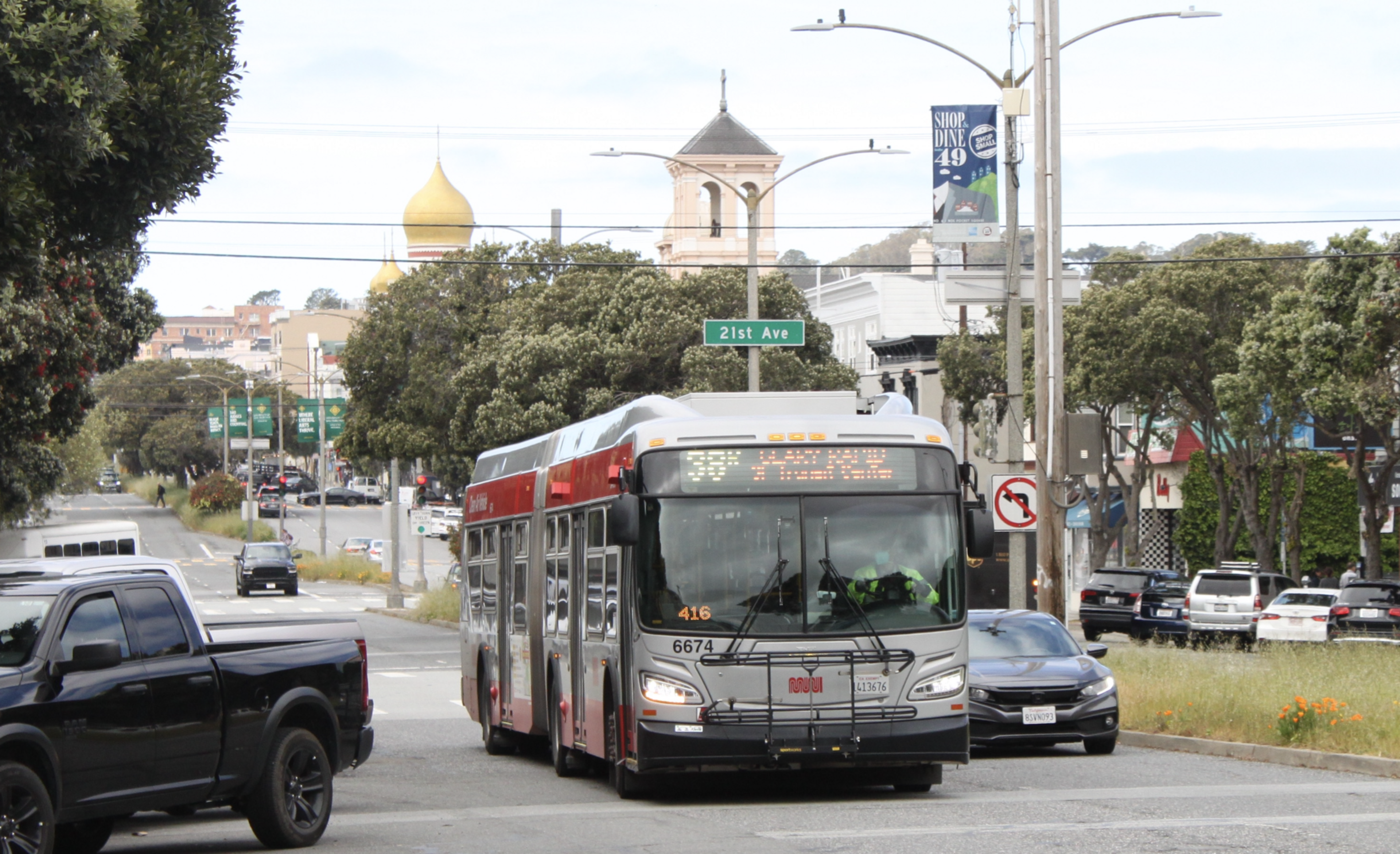 A red and silver city bus is driving down a street labeled 21st Ave, surrounded by cars, trees, and buildings with a golden dome and a bell tower visible in the background.