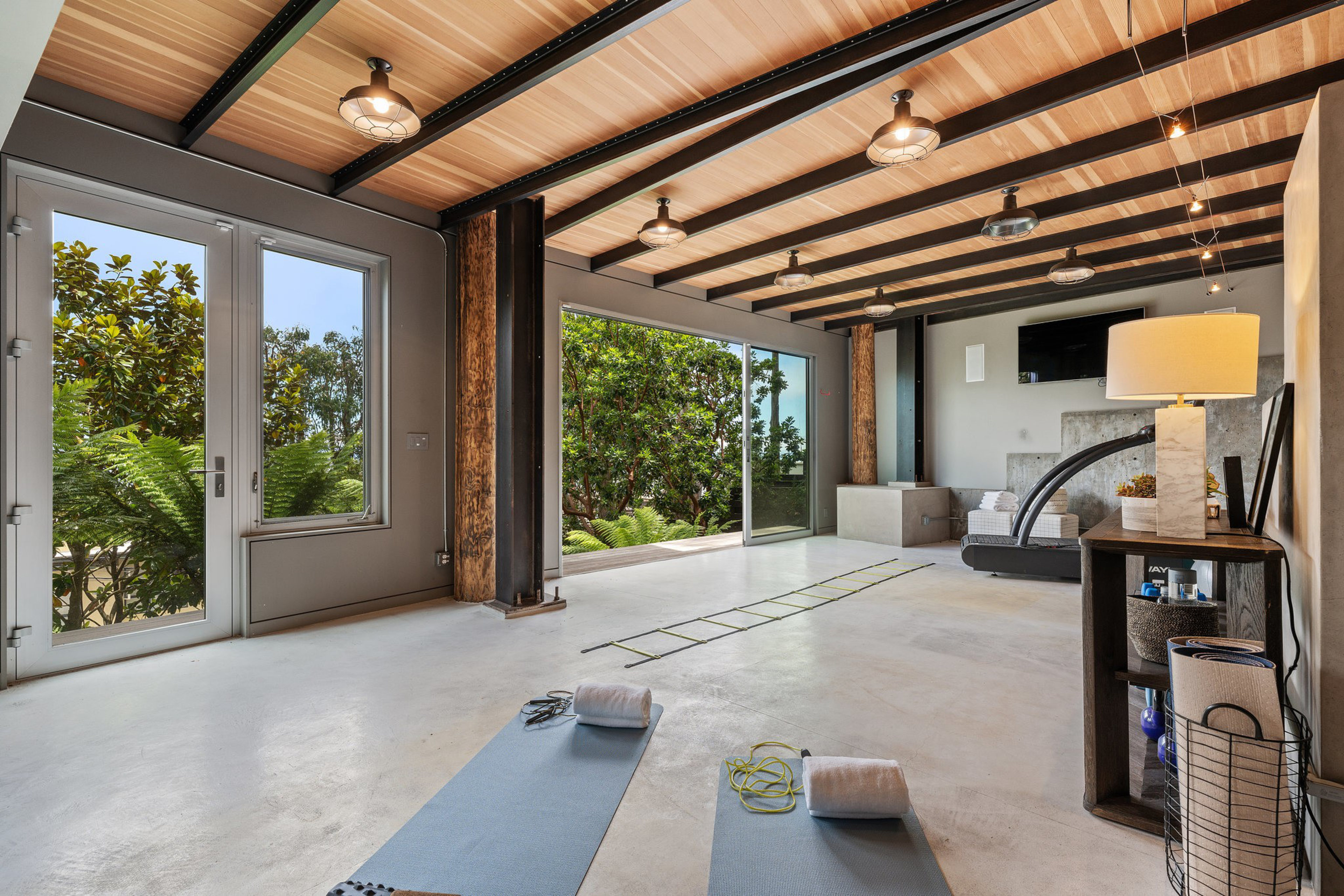 A spacious room with wooden ceiling beams, large sliding glass doors opening to greenery, two yoga mats with towels, and a cozy seating area with a TV.