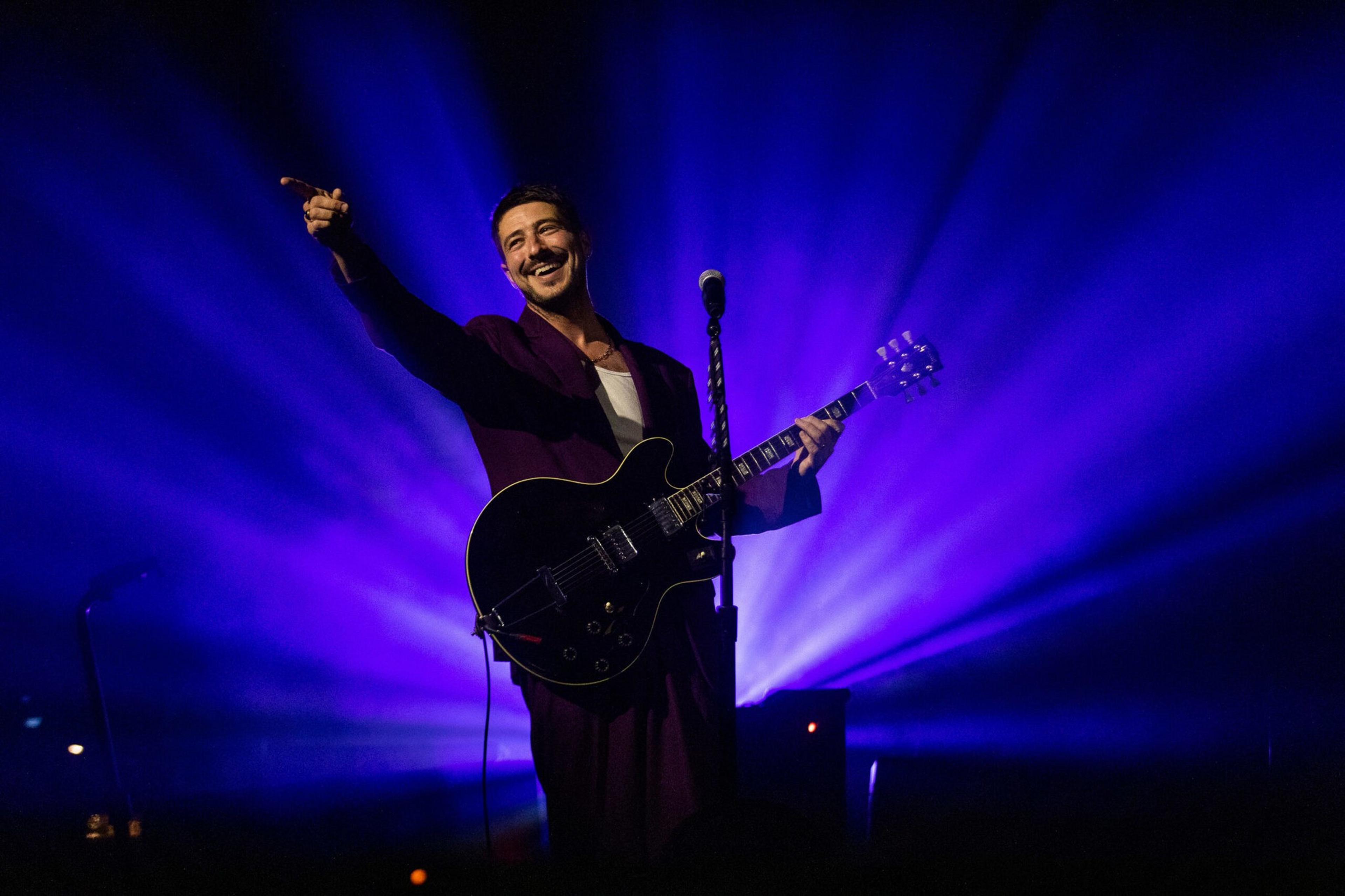 A smiling musician in a dark suit plays black electric guitar, points outward, and stands in front of a microphone with vibrant purple stage lights behind him.