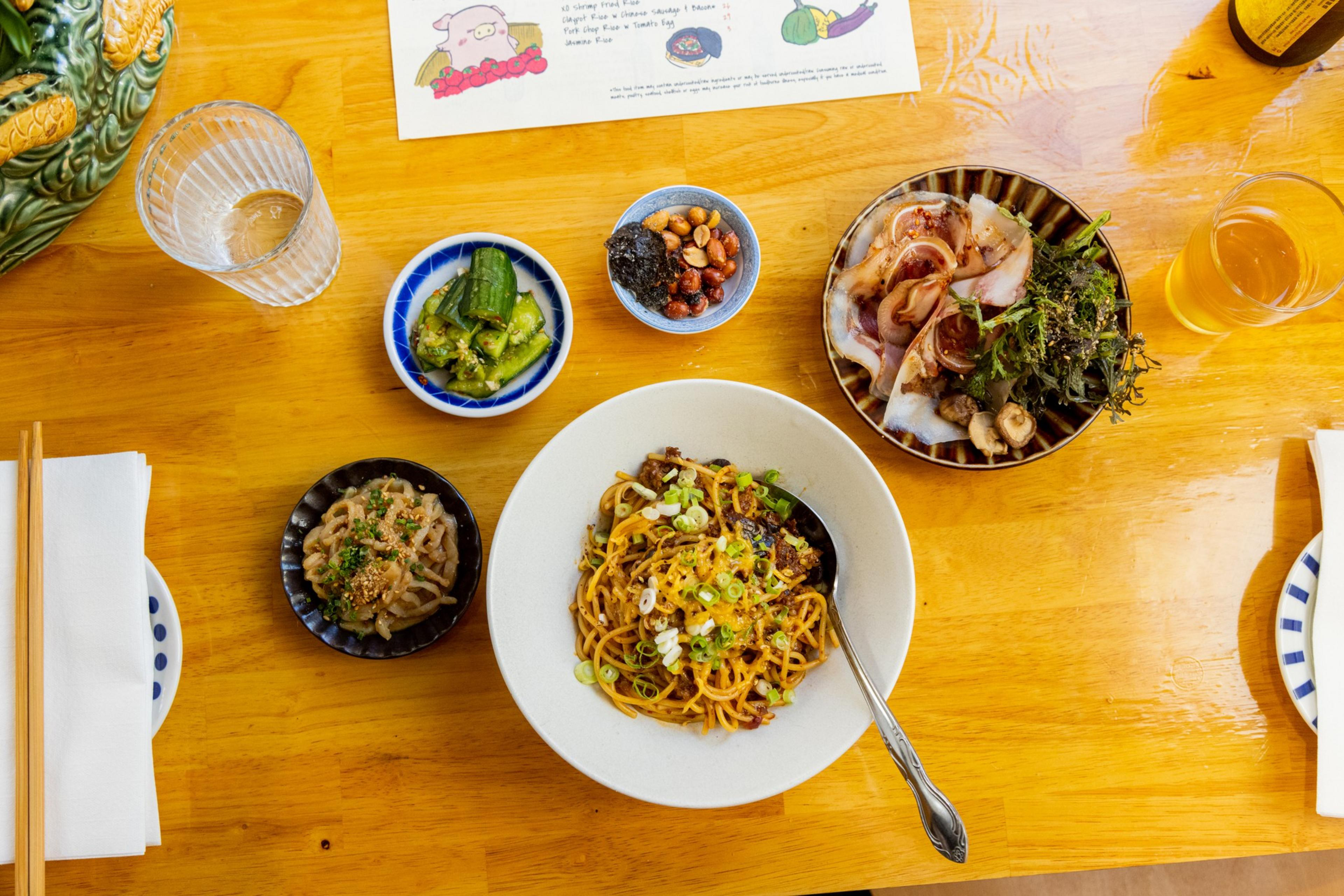 A wooden table holds a bowl of noodles with garnish, side dishes like cucumbers and seasoned mushrooms, assorted nuts, a glass of water, and another drink.