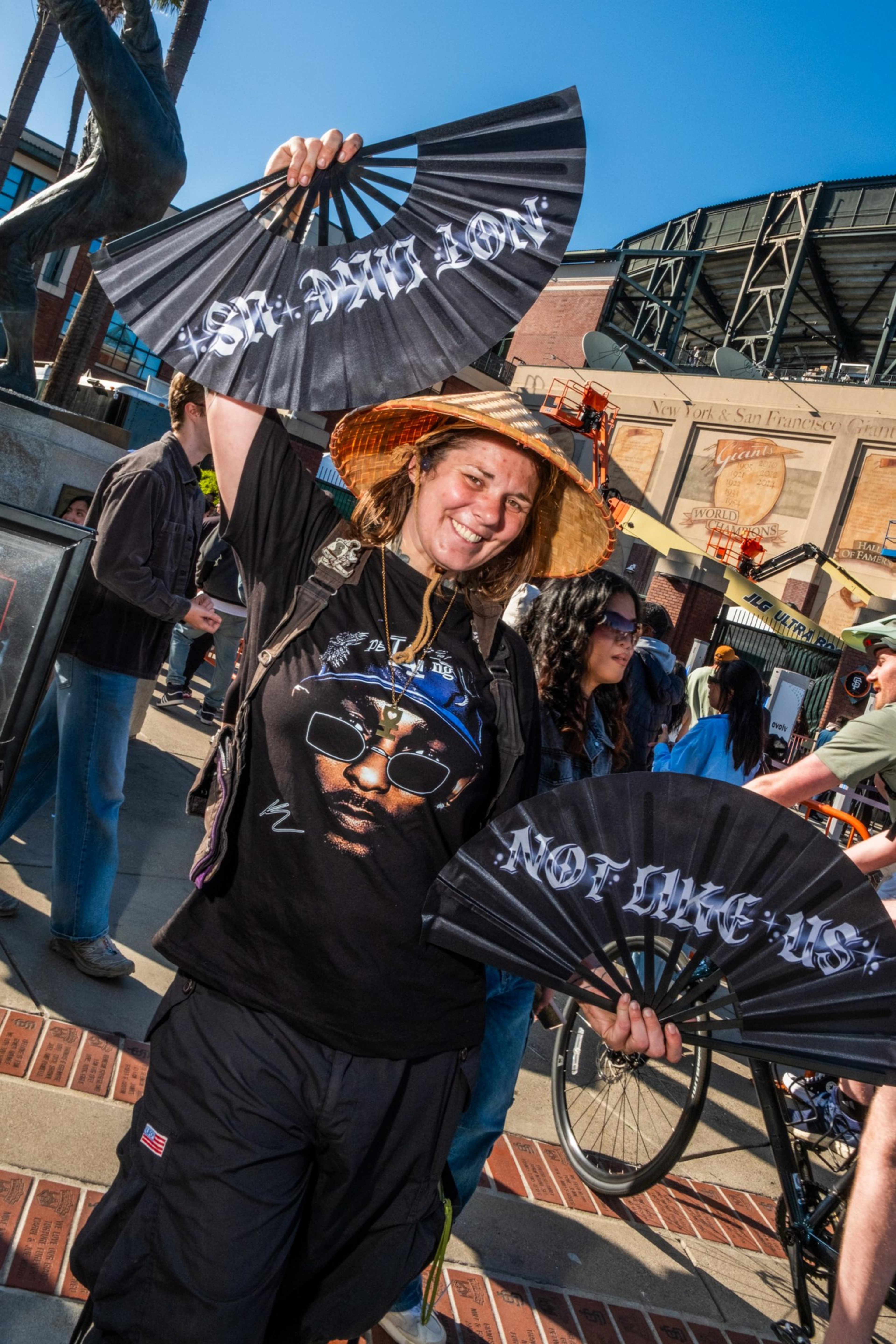 A smiling person wearing a straw hat holds two fans with text. They're in front of a building with a sports motif, and there's a statue and people nearby.