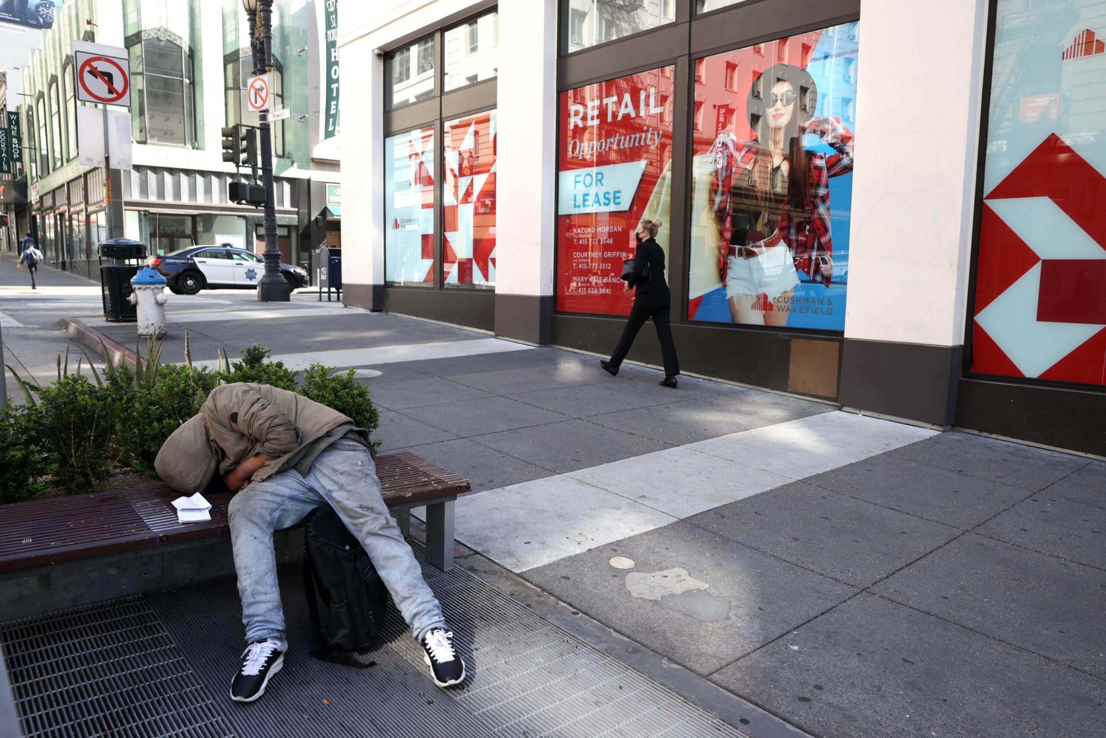 A person wearing a hooded jacket and jeans is slumped over a bench, while another person walks past a storefront with "For Lease" signs.