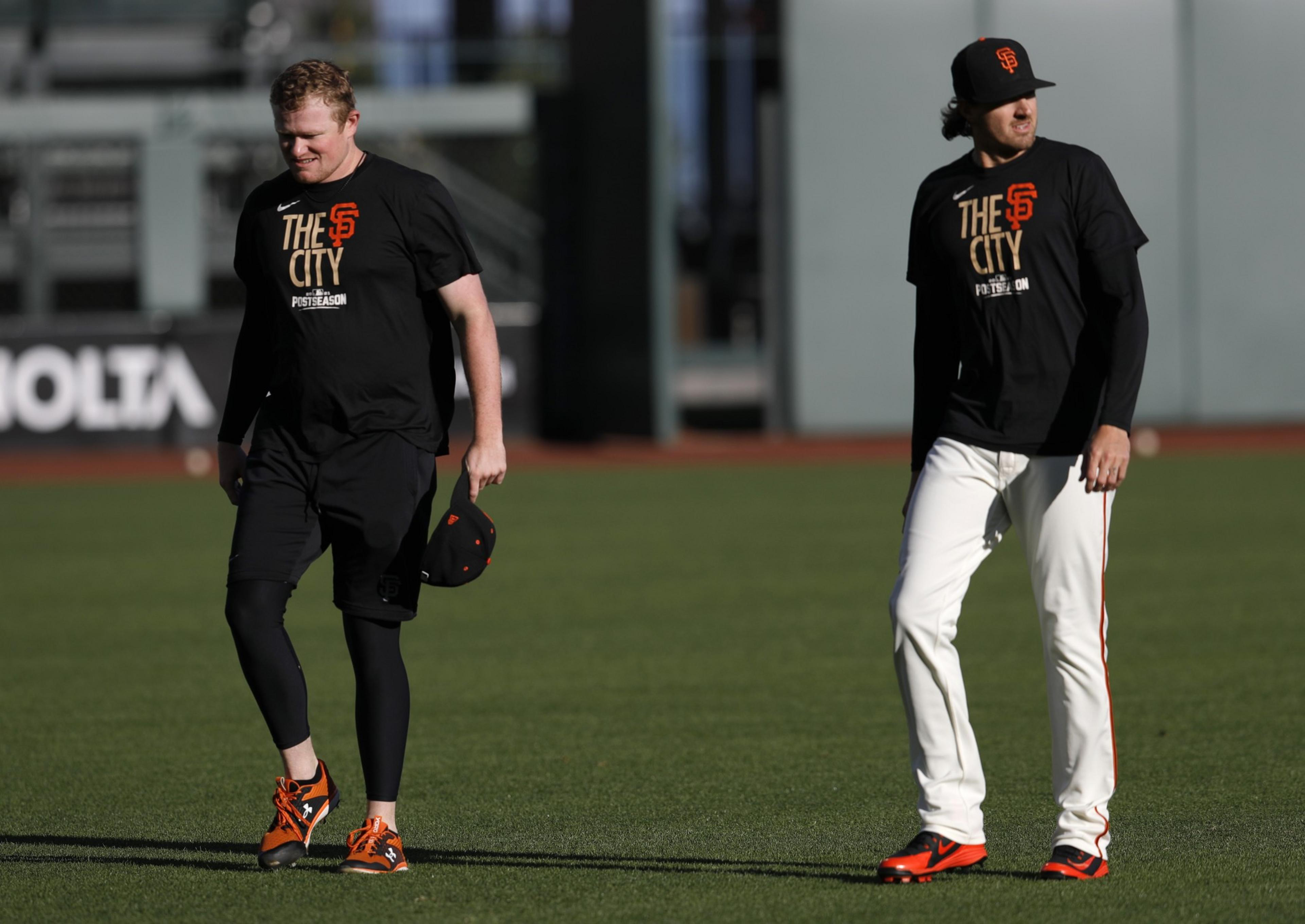 Two individuals stand on a baseball field wearing black shirts with &quot;THE CITY&quot; and team logos. One wears black pants, the other white, both have orange shoes.