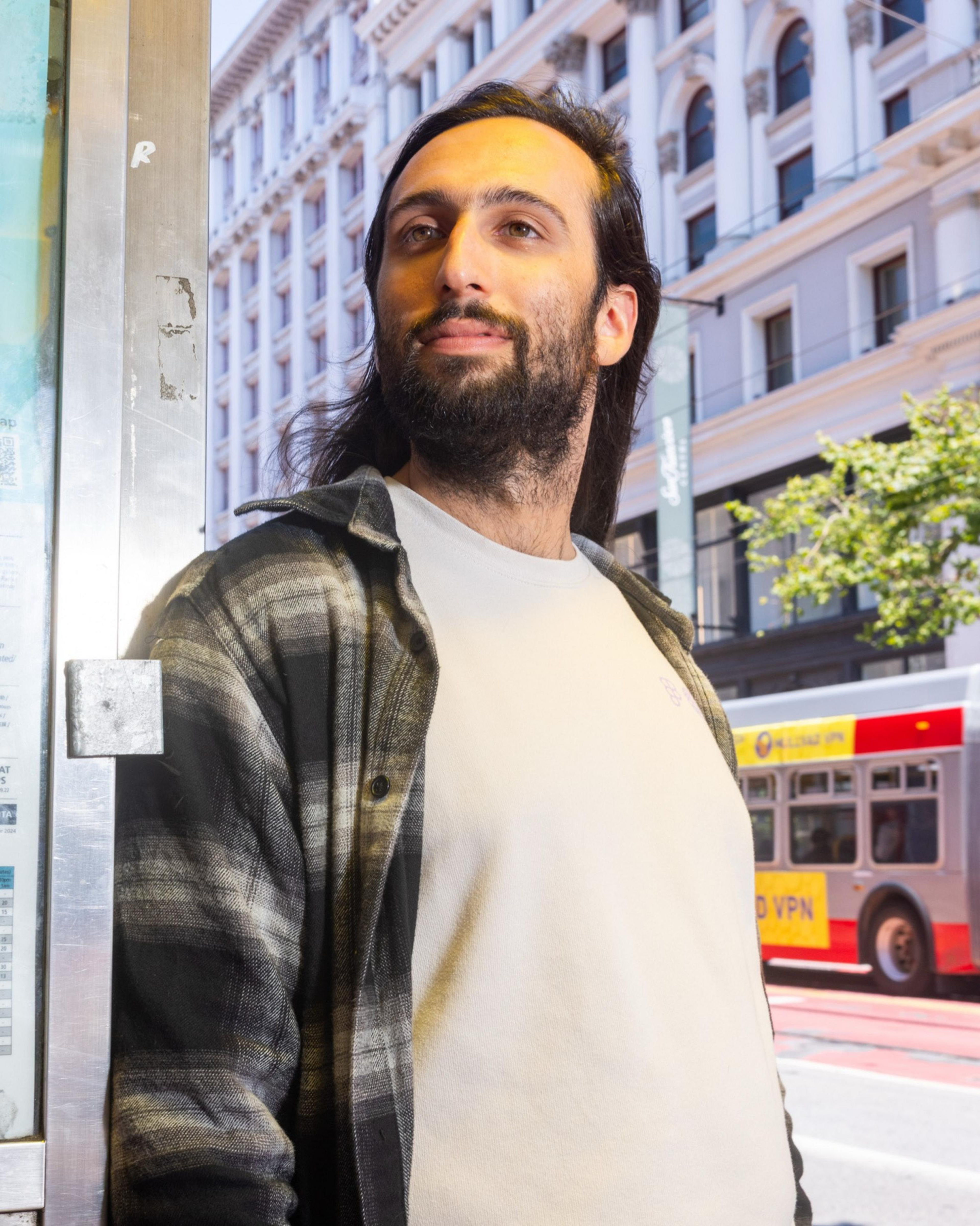 A person with long dark hair and a beard stands by a structure, wearing a plaid shirt over a white tee. A building and bus are in the sunny background.