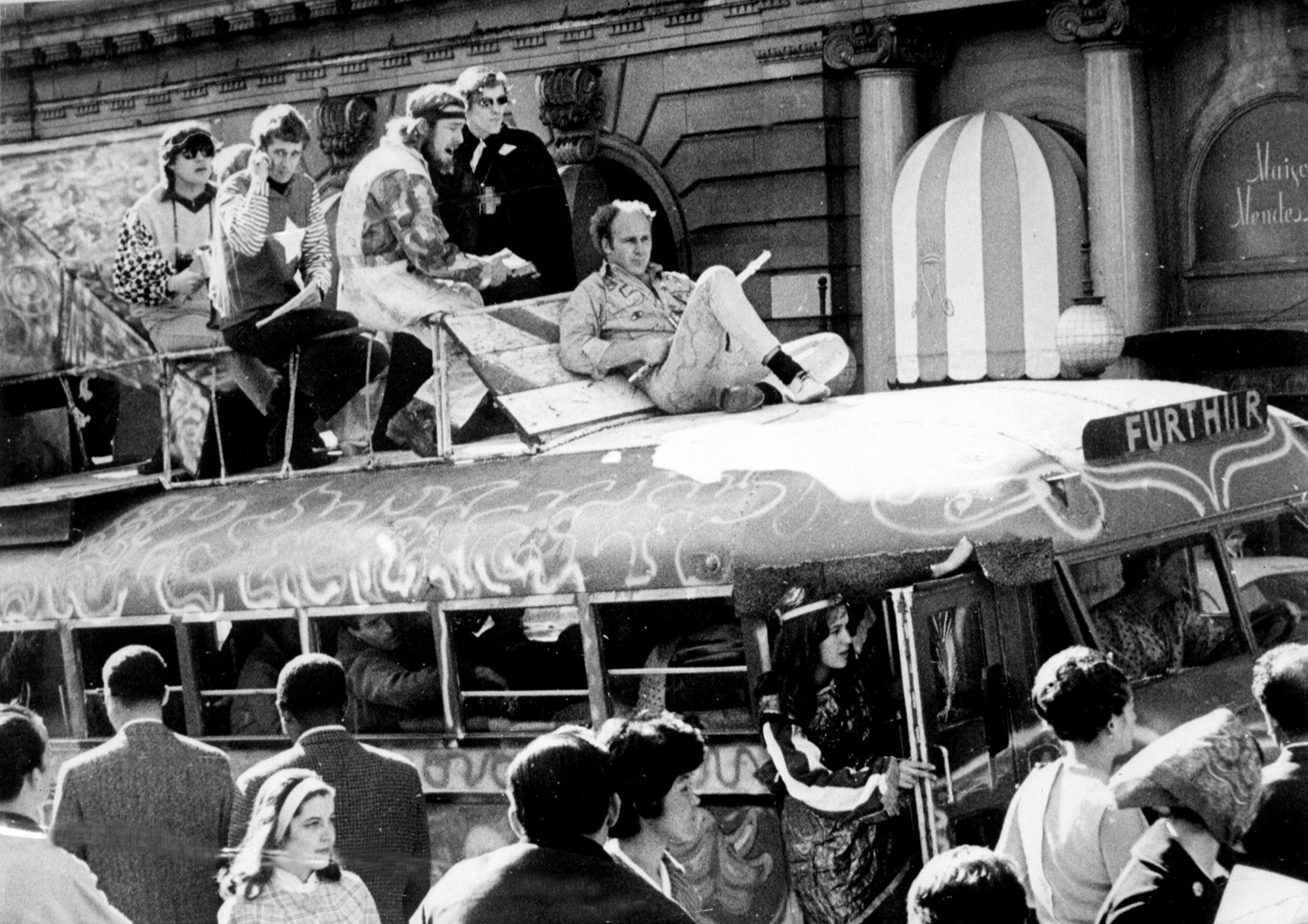 Several people sit and stand on top of a creatively painted bus labeled "FURTHER," while a crowd gathers below near a building with columns.