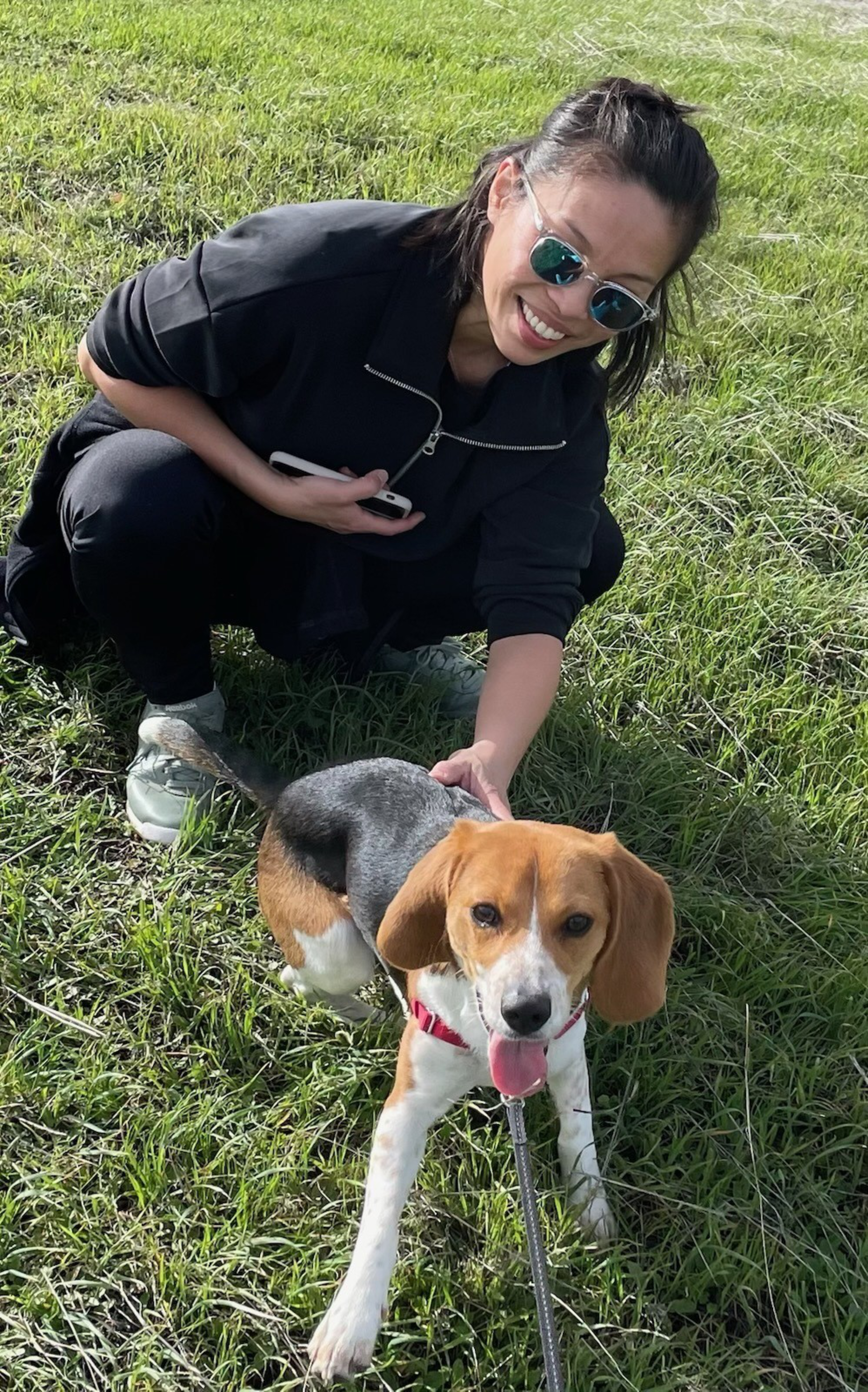 A woman wearing sunglasses crouches on grass, smiling at the camera. She holds a phone and pets a happy beagle with a red harness beside her.