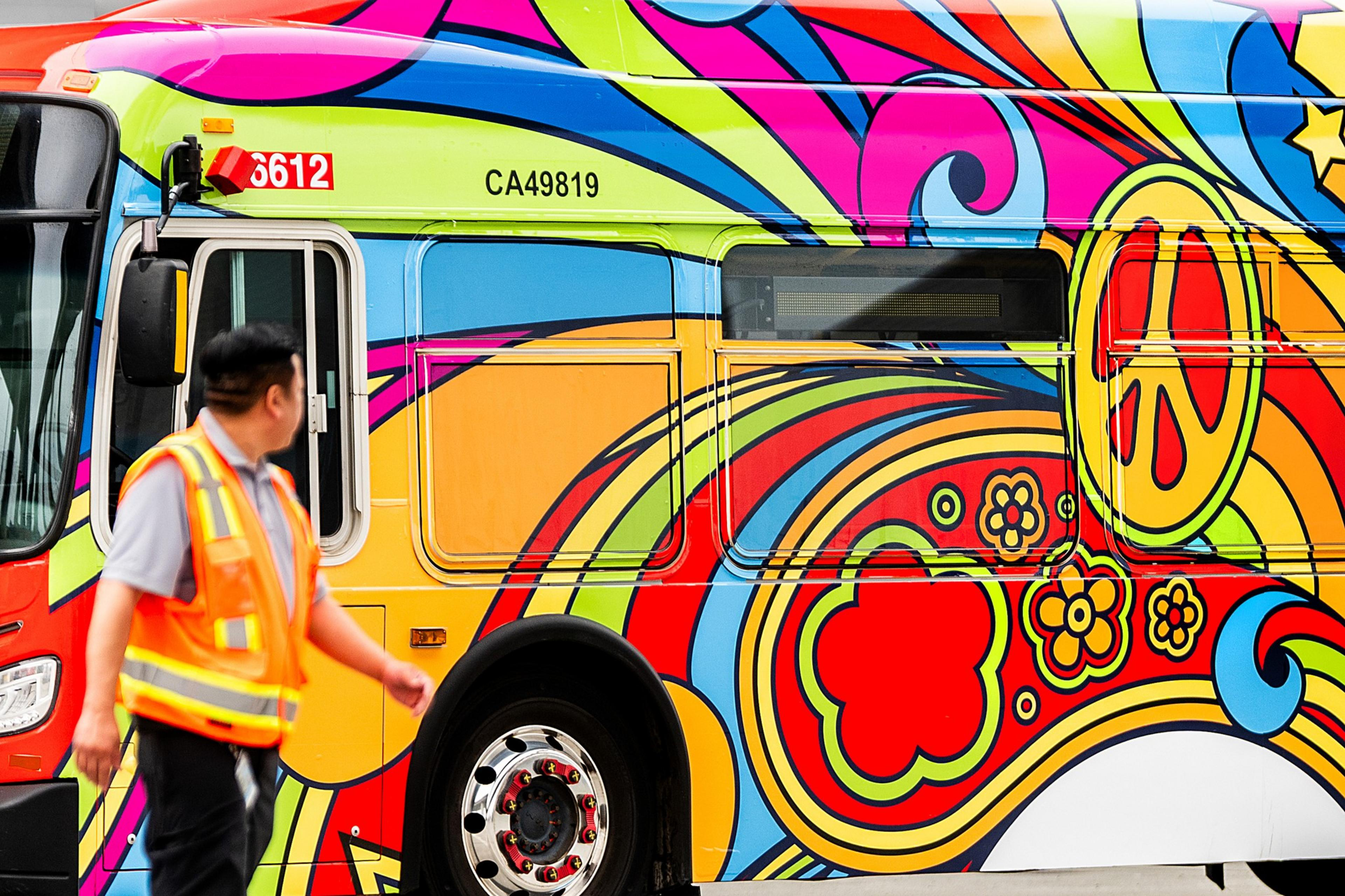 A vibrantly painted bus features psychedelic patterns with swirls, flowers, and peace signs. A person in an orange safety vest walks by.
