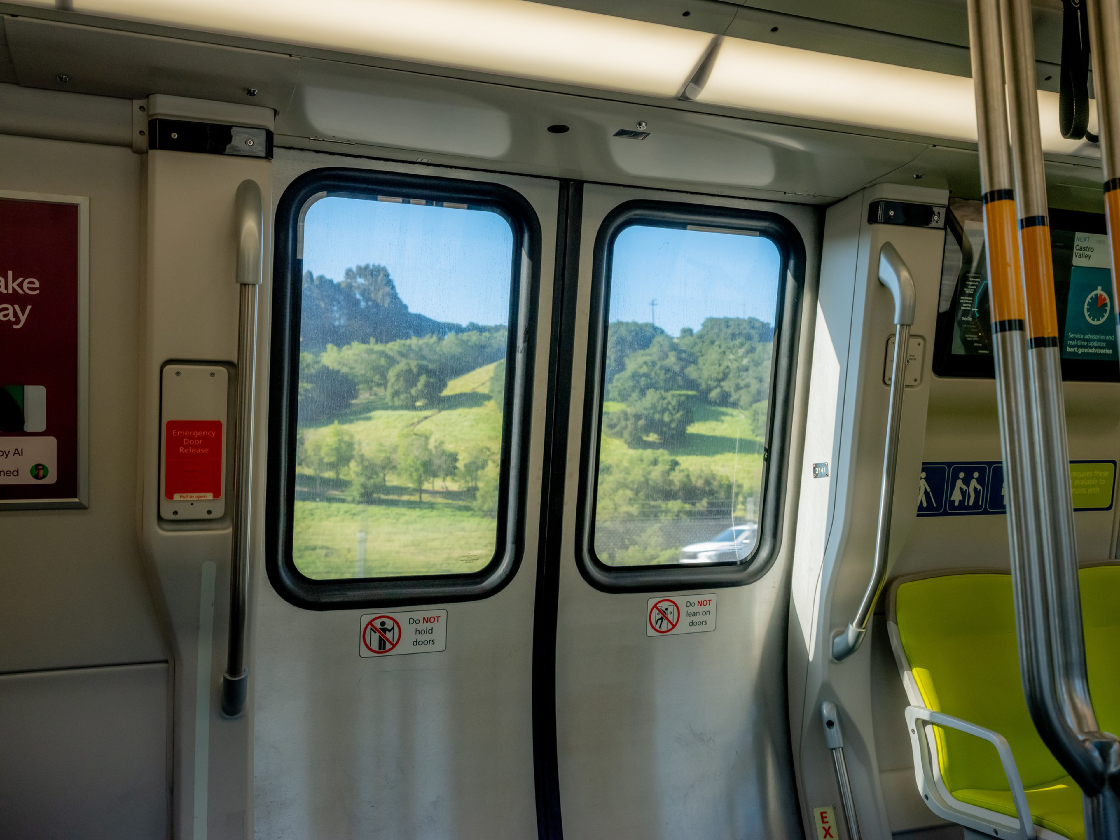 Train doors with windows revealing a sunny landscape of green hills and trees outside, nearby yellow seats and metal grab bars inside.