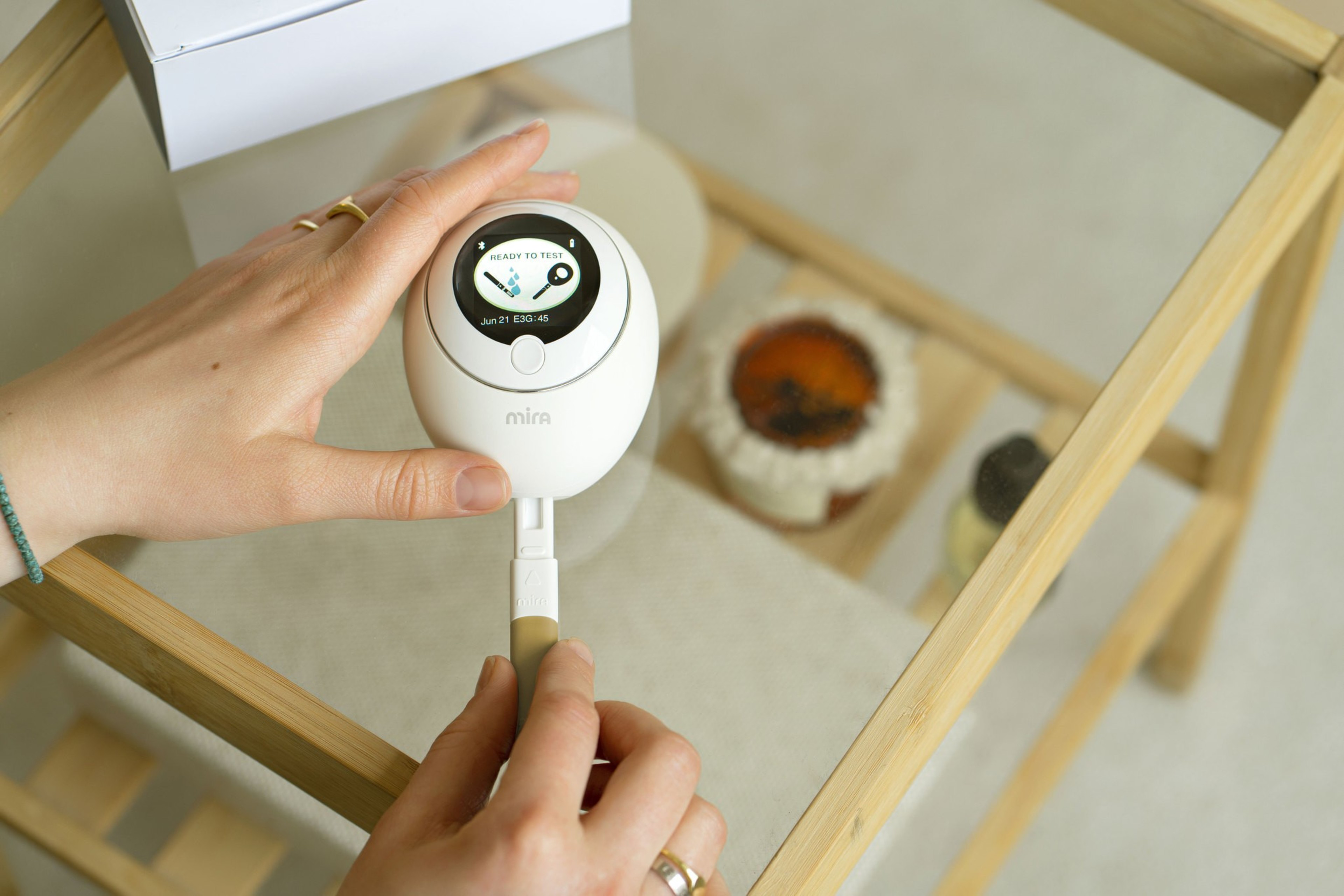 A person holds a handheld device with a circular screen showing "Ready to test." The device is labeled "Mira," and it rests on a glass surface with jars below.