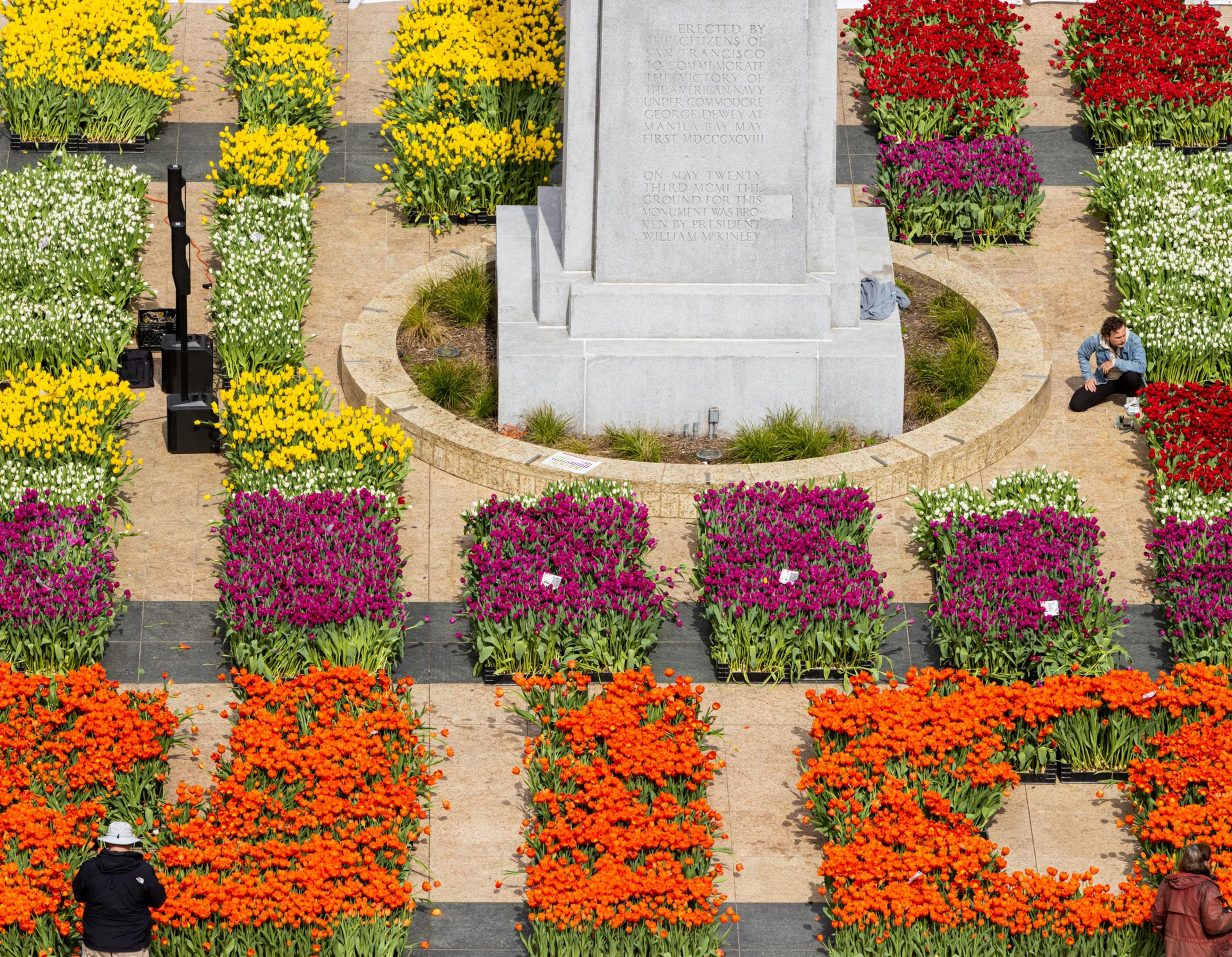 A vibrant display of red, orange, yellow, and purple flowers surrounds a monument. Two people crouch near the flowers, likely tending them.