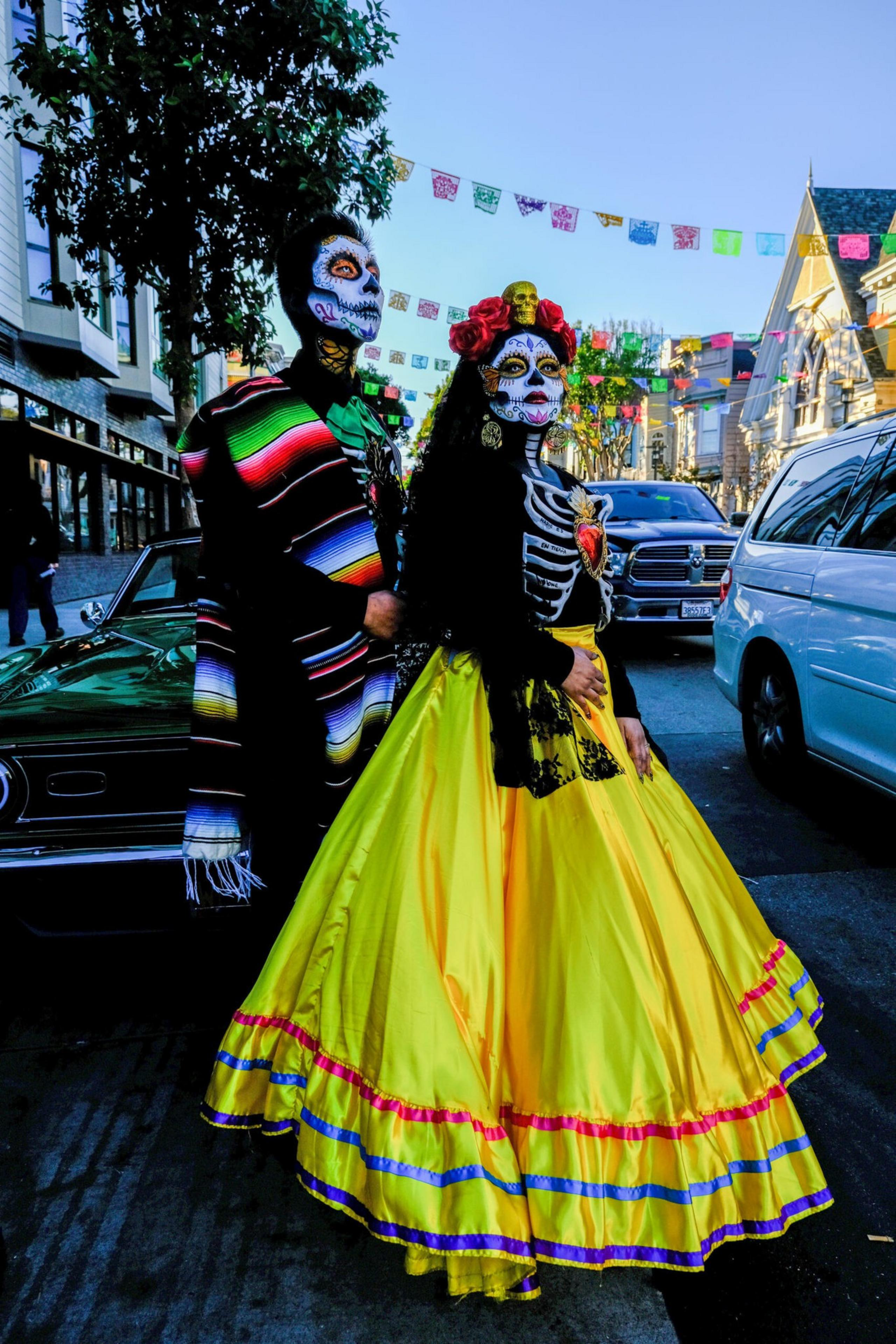 A couple stands dressed in vibrant Day of the Dead costumes with skull face paint; the woman wears a bright yellow skirt with red and blue trim.
