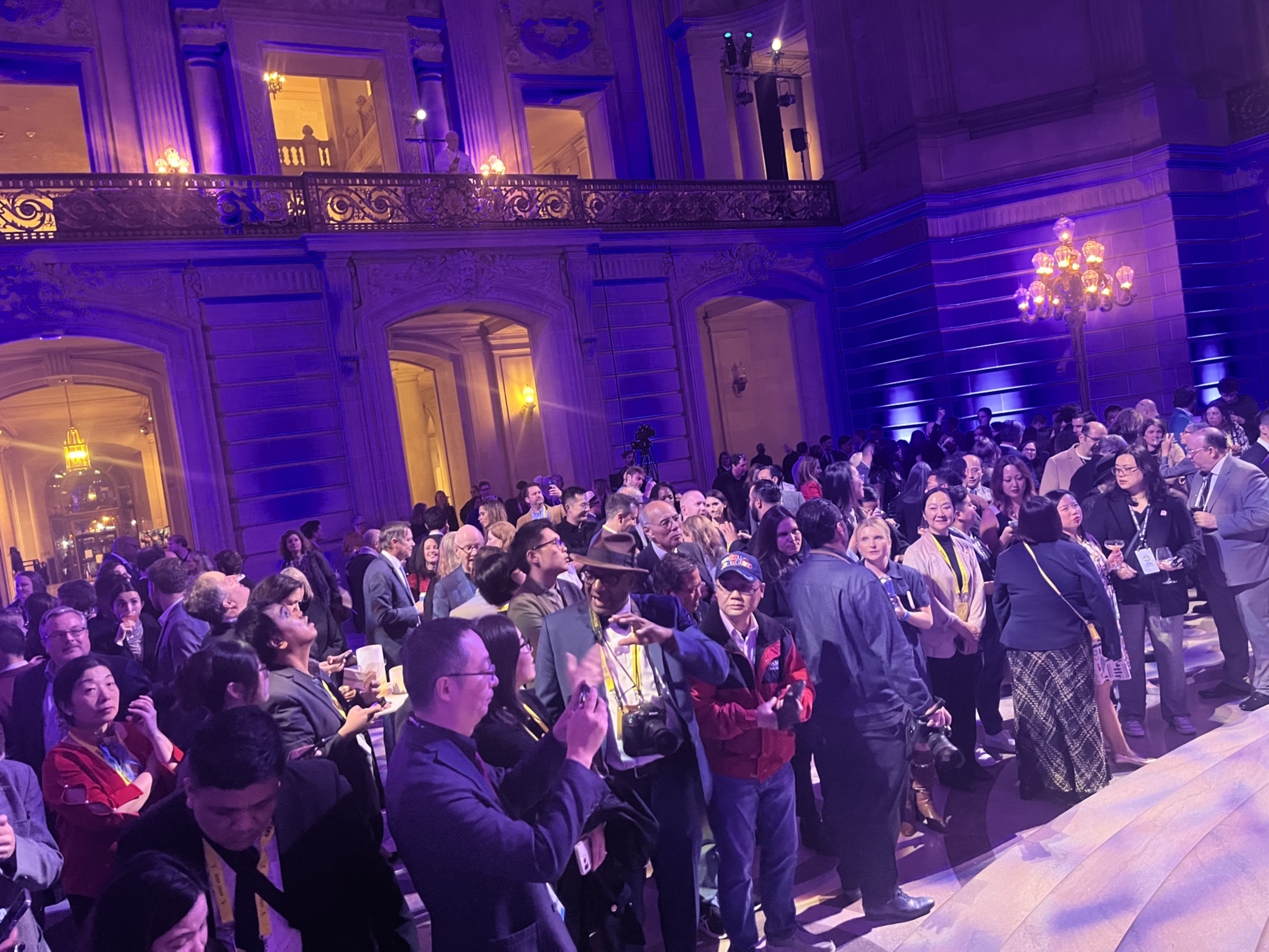 A group of journalists stand in a formal rotunda of a government building waiting for speeches to start.