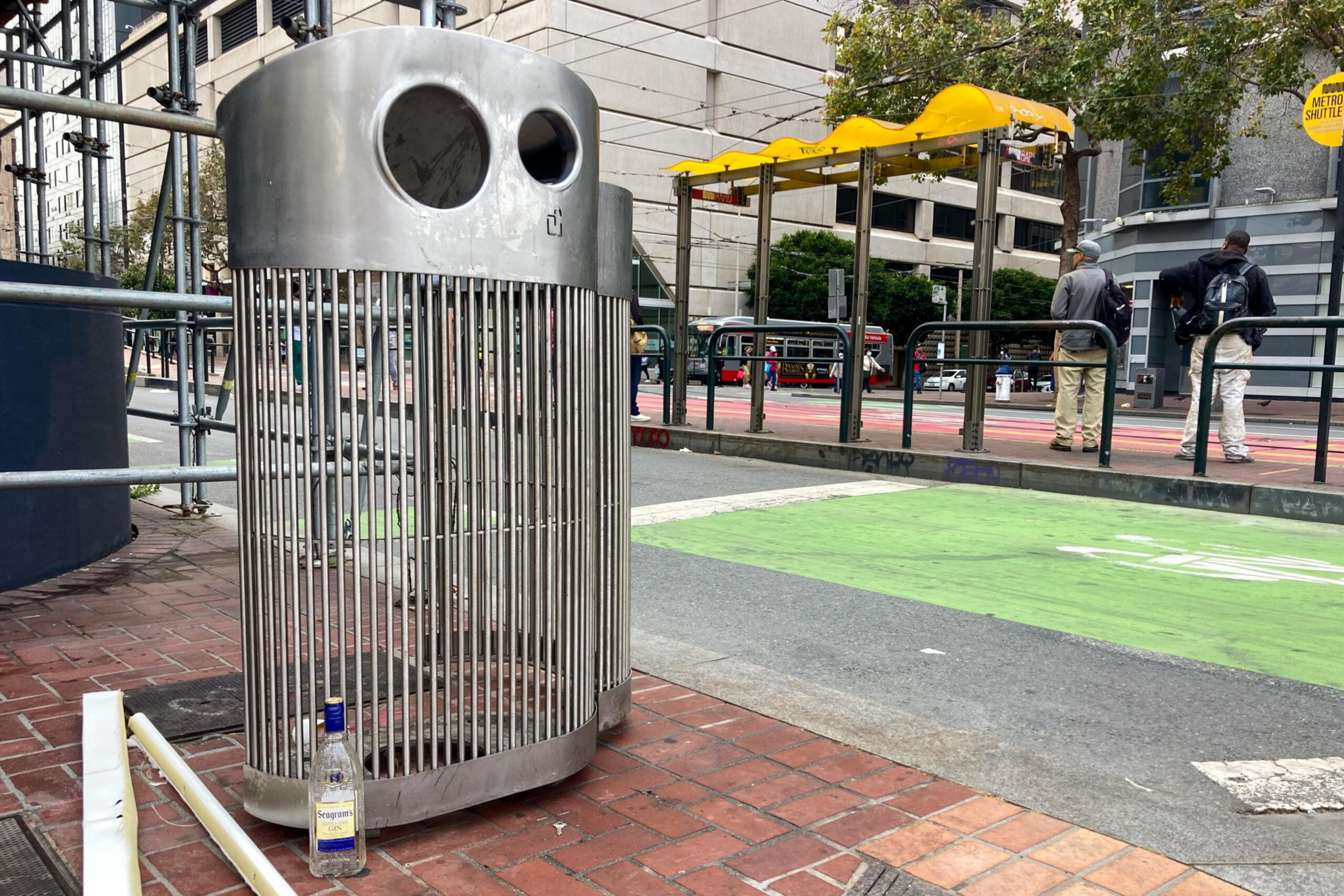 A cylindrical metal trash bin is on a city sidewalk, next to which is an empty alcohol bottle. In the background, people wait at a bus or tram stop.