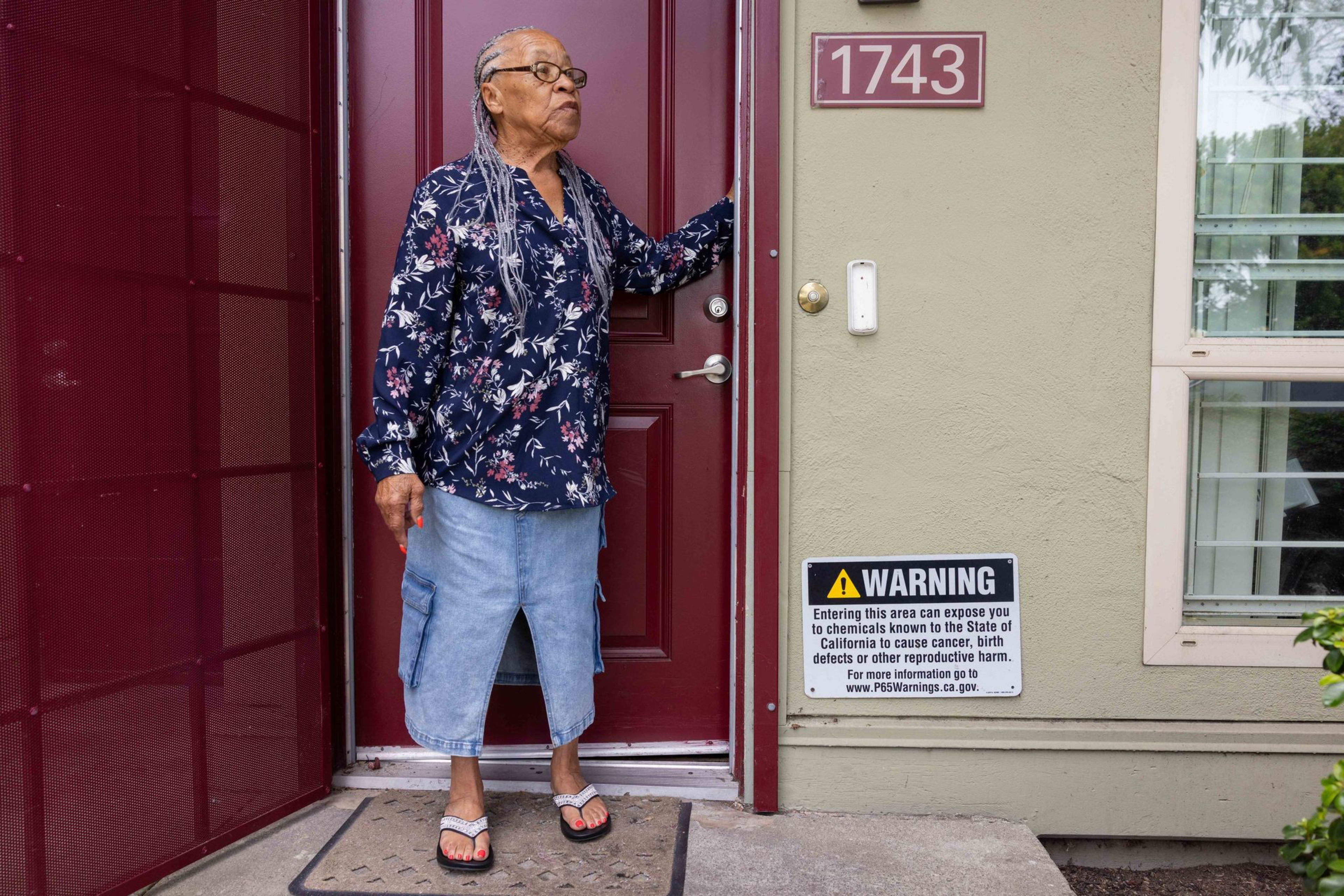 An elderly woman with gray braids and glasses stands at a maroon door, wearing a floral blouse, denim skirt, and sandals, next to a warning sign and house number 1743.