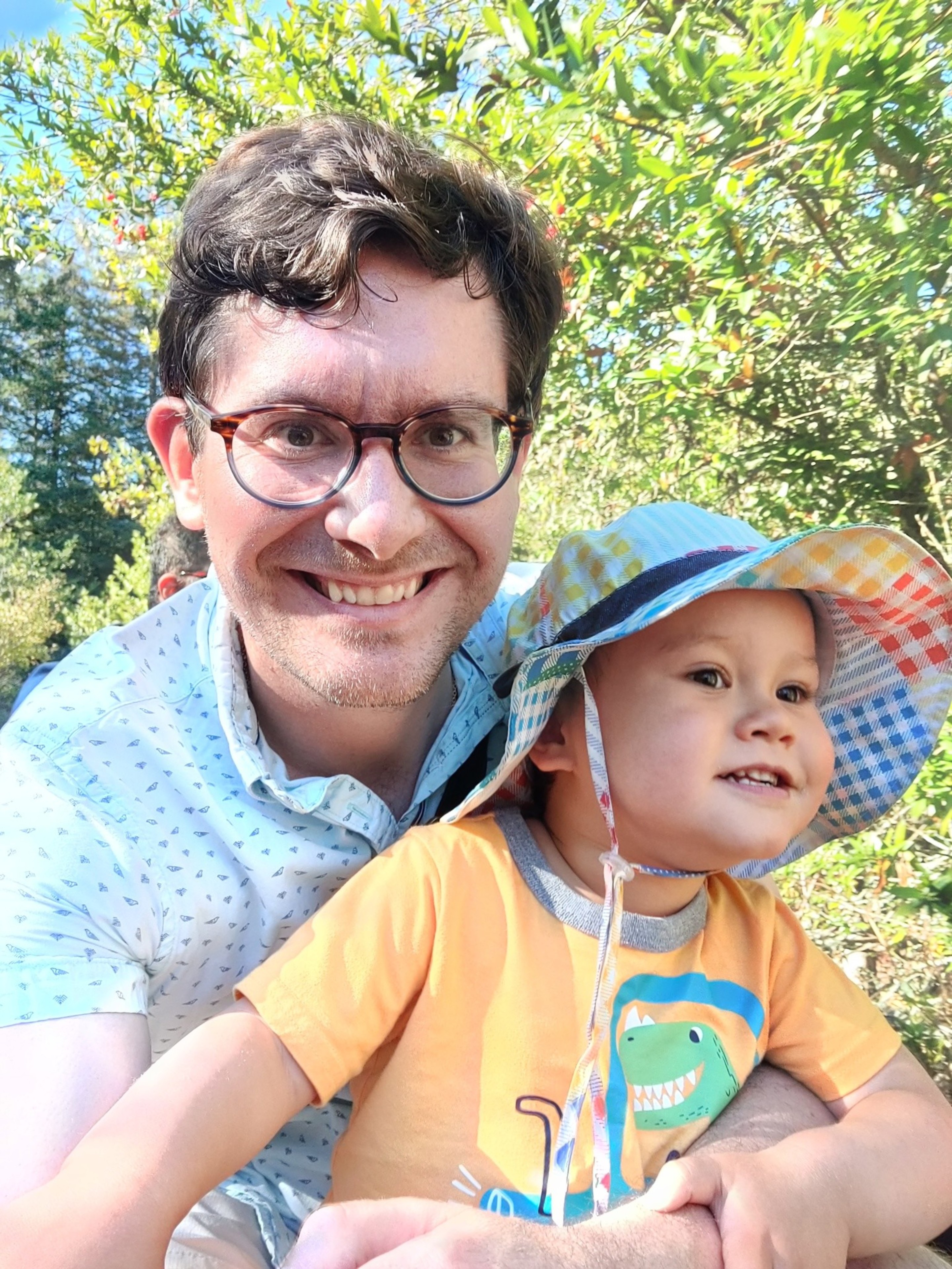 A smiling man with glasses and a child wearing a colorful sunhat and dinosaur-themed shirt pose outdoors with green foliage in the background.