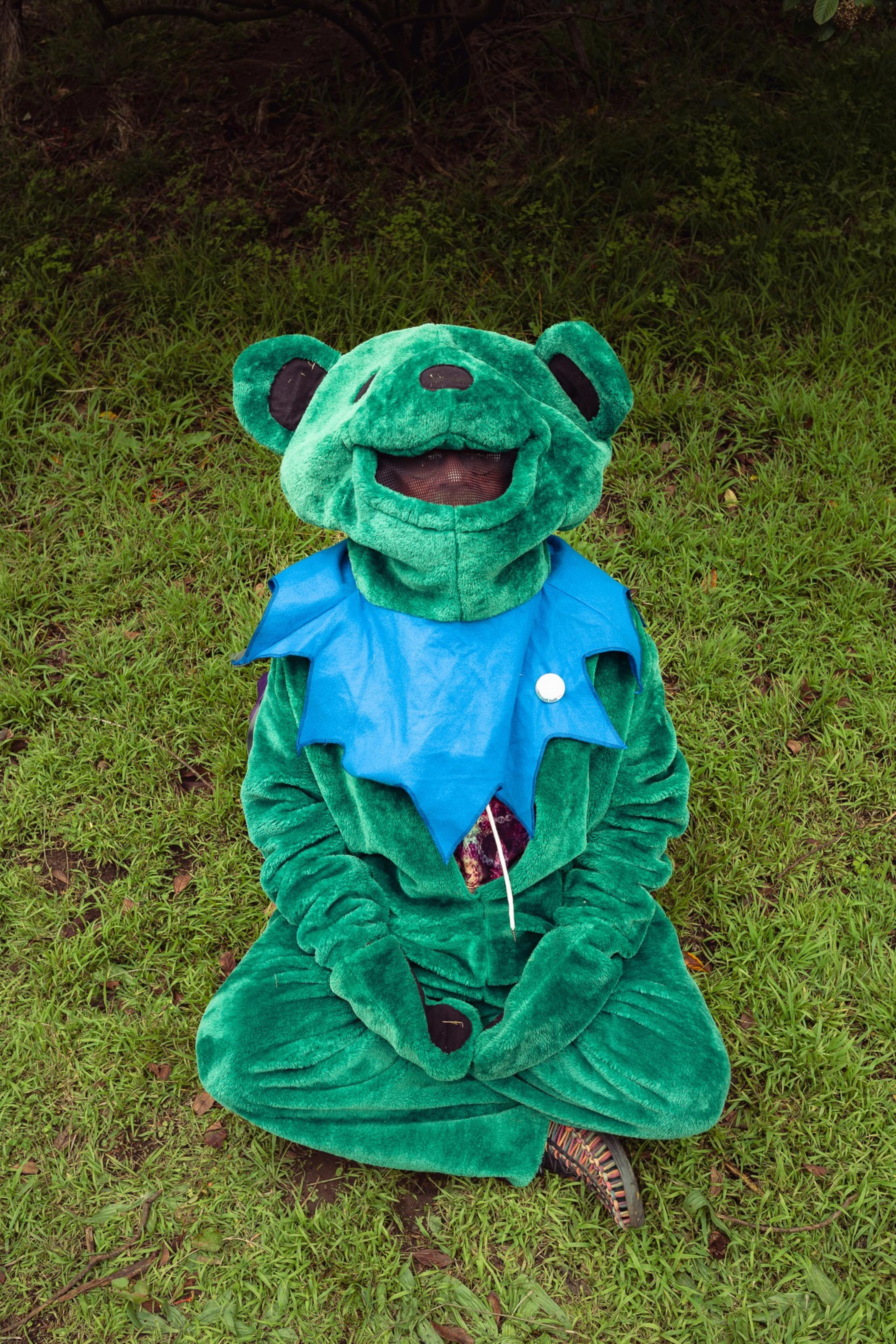 A person dressed in a green bear costume with a blue collar sits cross-legged on grass against a natural, leafy background.