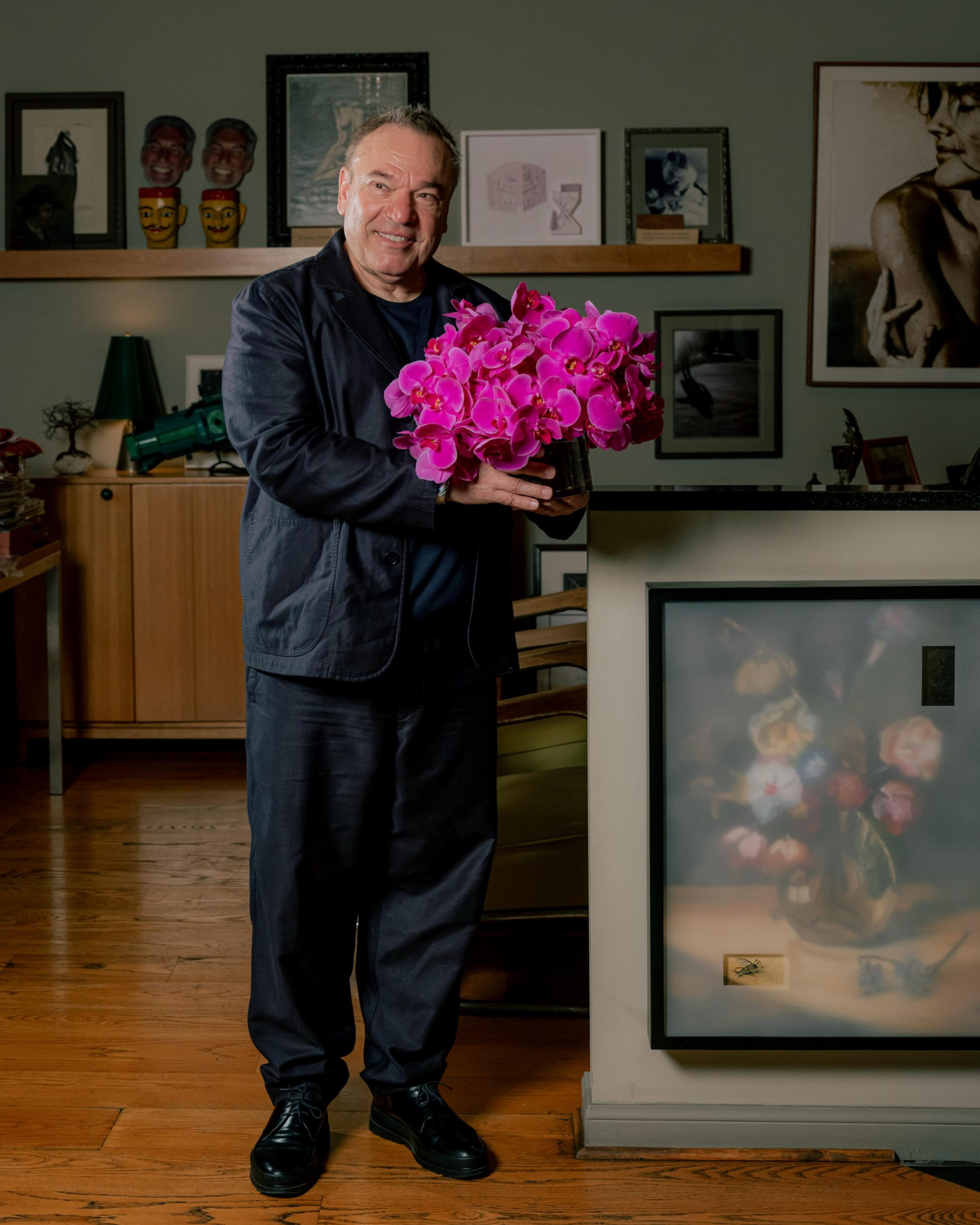 A man in dark clothing smiles while holding a vibrant bouquet of pink orchids in a room with wooden floors and framed artwork on the walls.