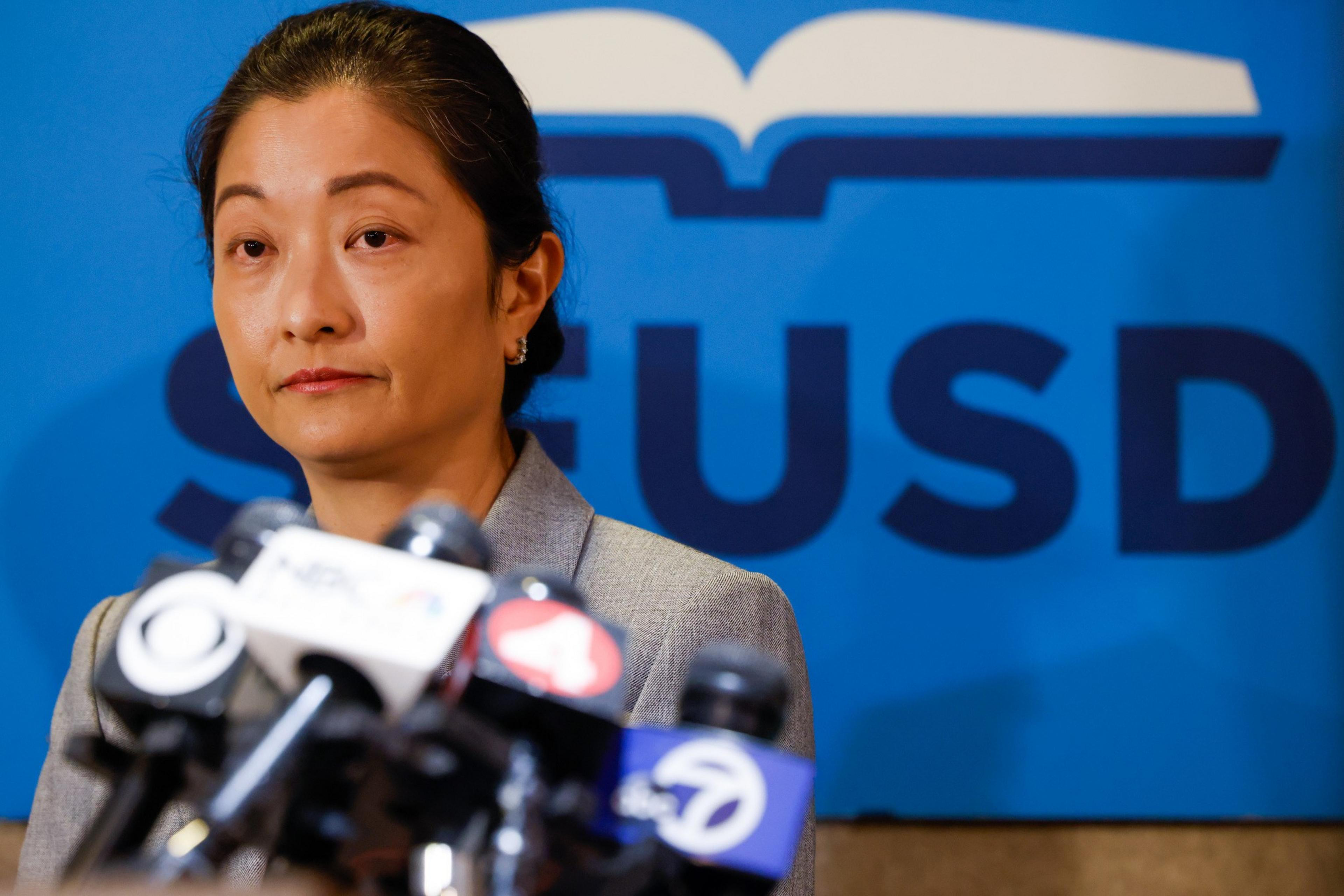 A person in a gray jacket stands before multiple microphones. The backdrop is blue with "SFUSD" and an open book logo visible.