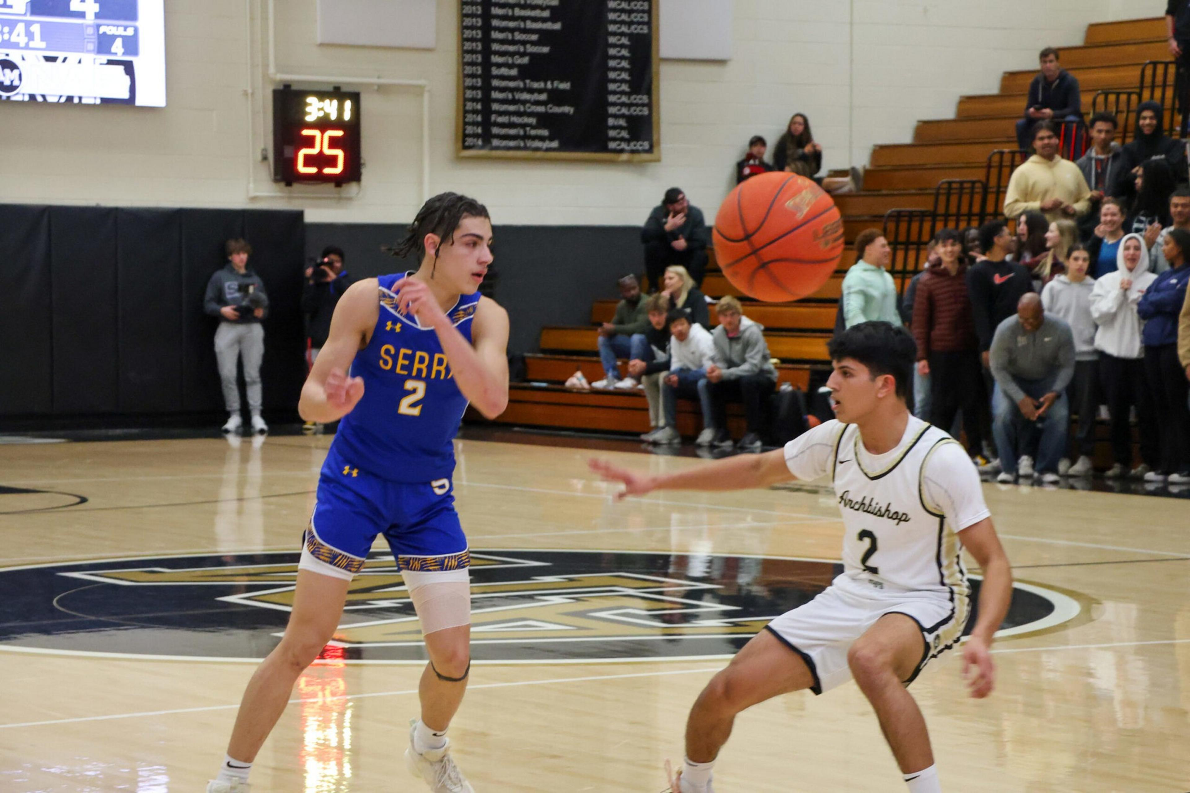 A basketball player in a blue jersey passes the ball while a player in a white jersey defends, with spectators watching from the bleachers.