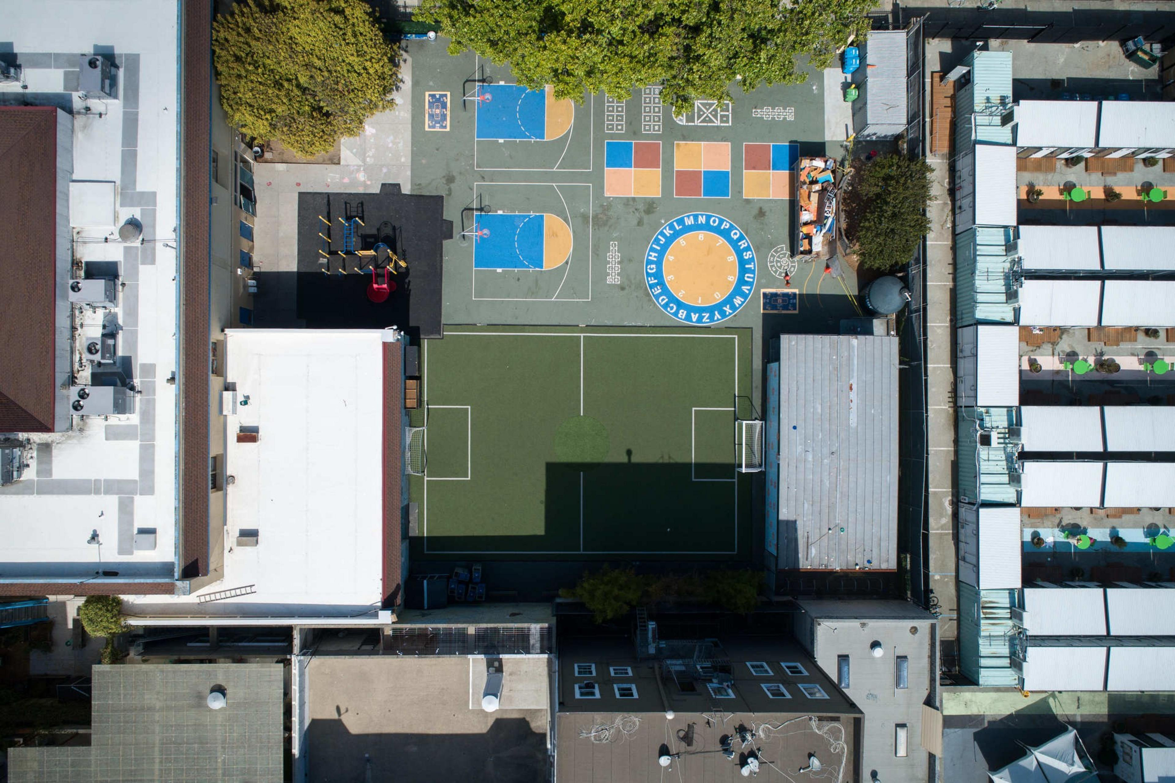 The image shows an aerial view of a colorful school playground with a basketball court, playground equipment, a small soccer field, and a seating area.