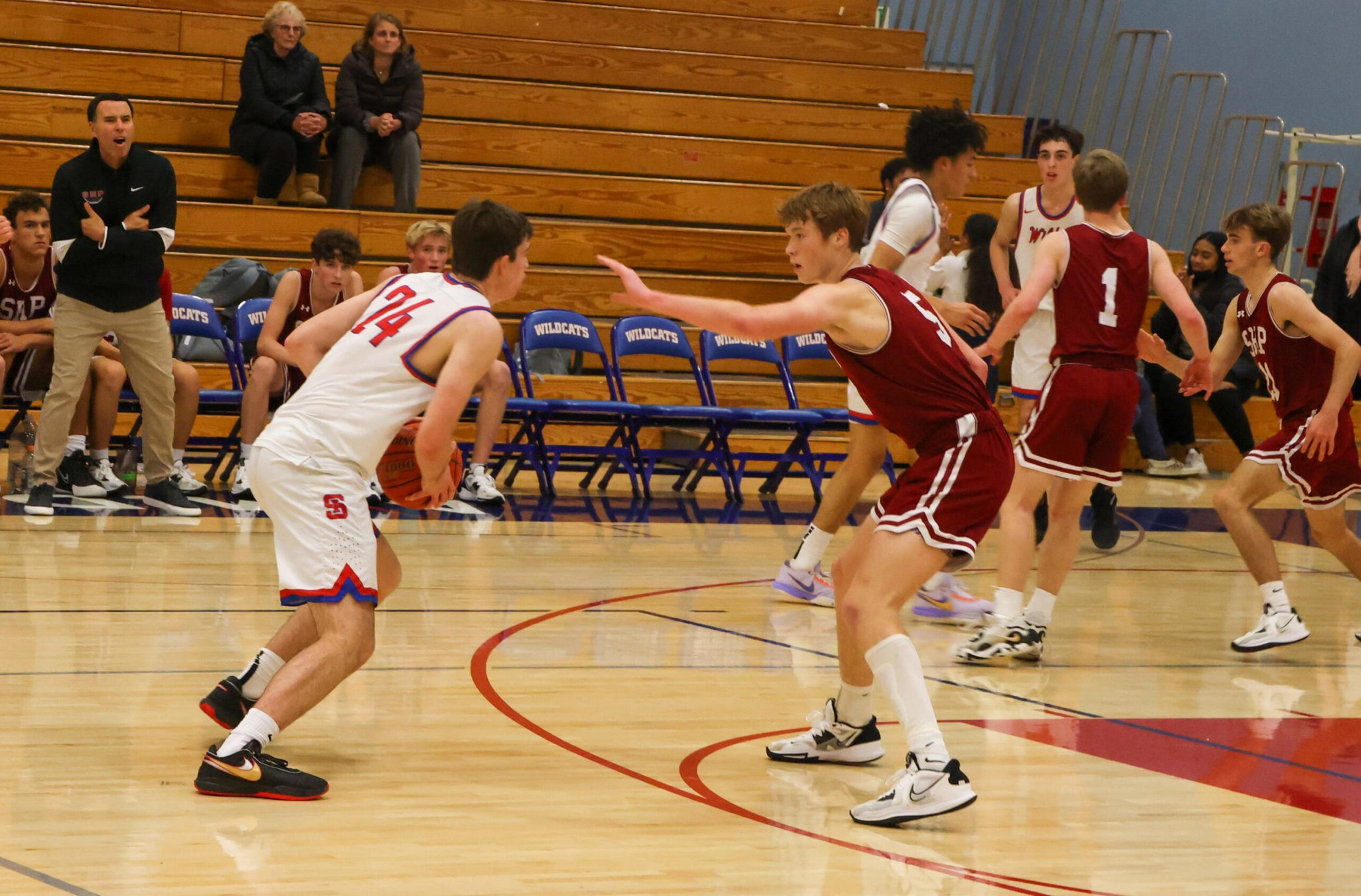 A basketball player in white prepares to pass while being guarded by a player in maroon in a gym with empty blue chairs labeled "WILDCATS."