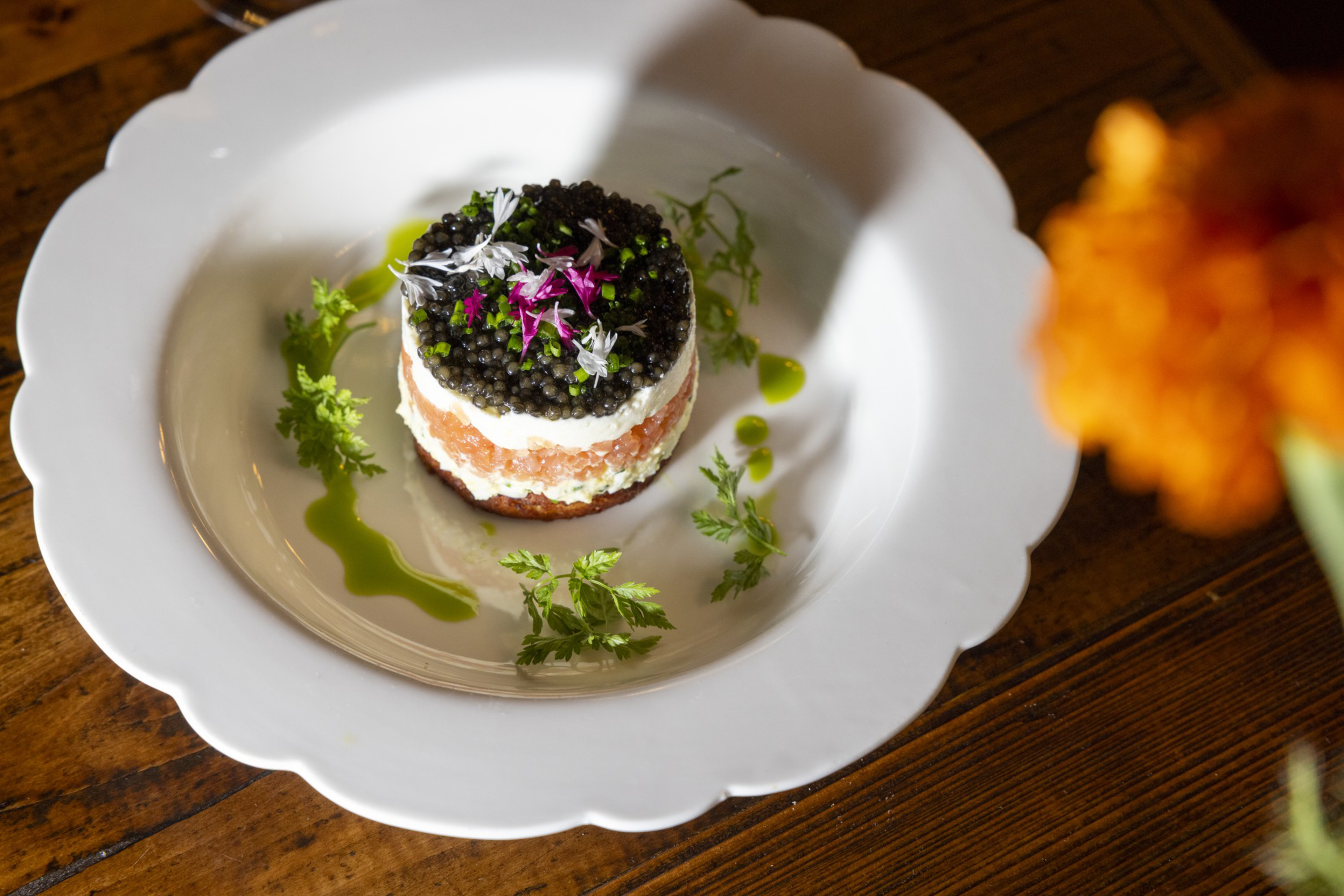 A gourmet dish sits on a white scalloped plate, featuring a layered cylinder of salmon, cream, and caviar, garnished with herbs, edible flowers, and green sauce.