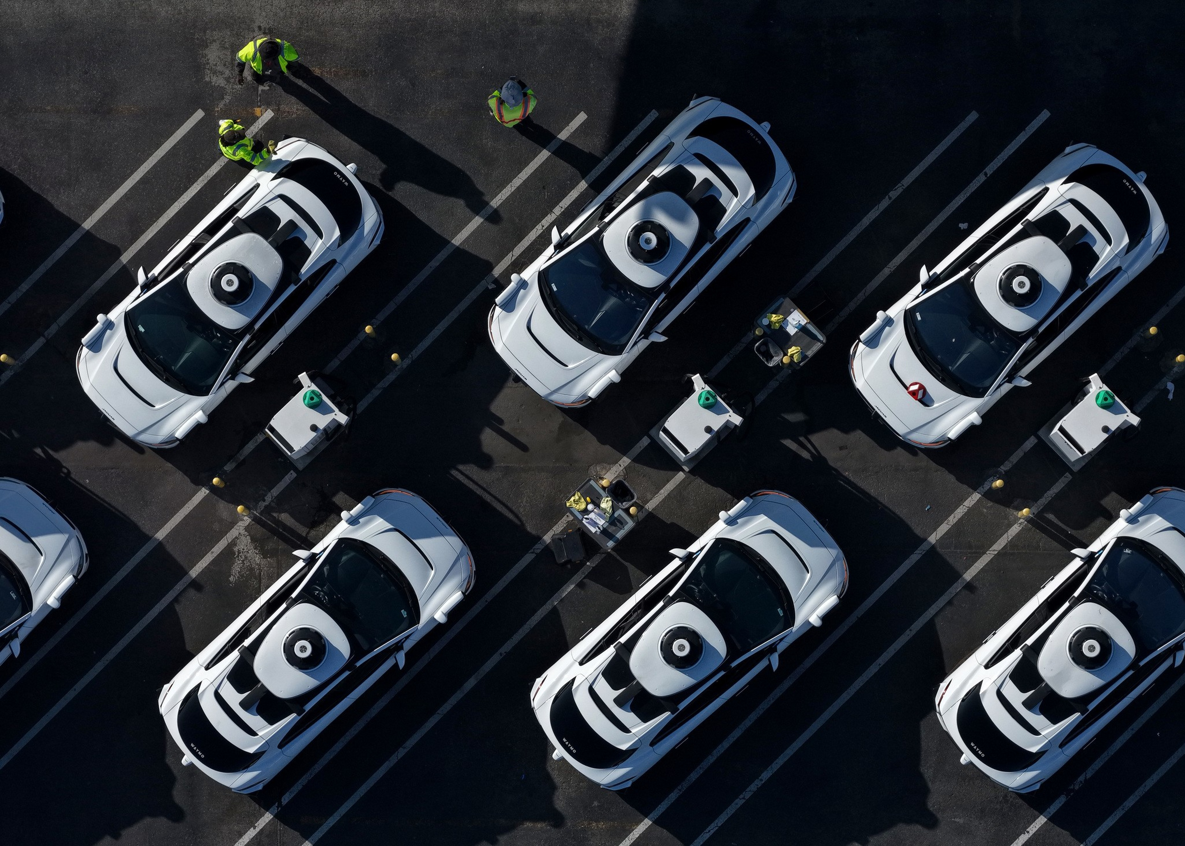 Six white self-driving cars are parked in a lot, with three workers in yellow vests standing and interacting near them.