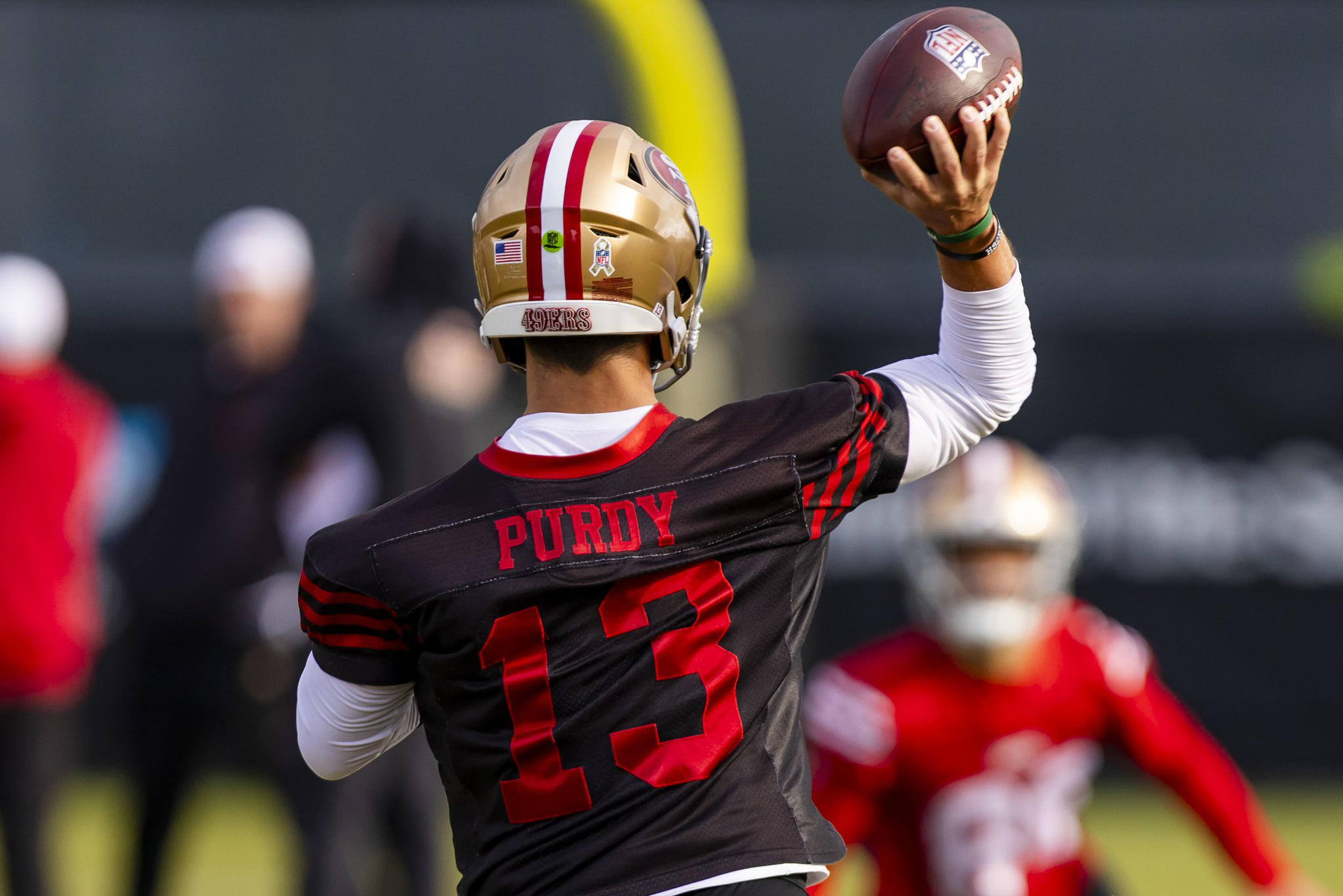 A football player wearing a black jersey with "Purdy" and number 13 prepares to throw a football during practice, with another player blurred in the background.