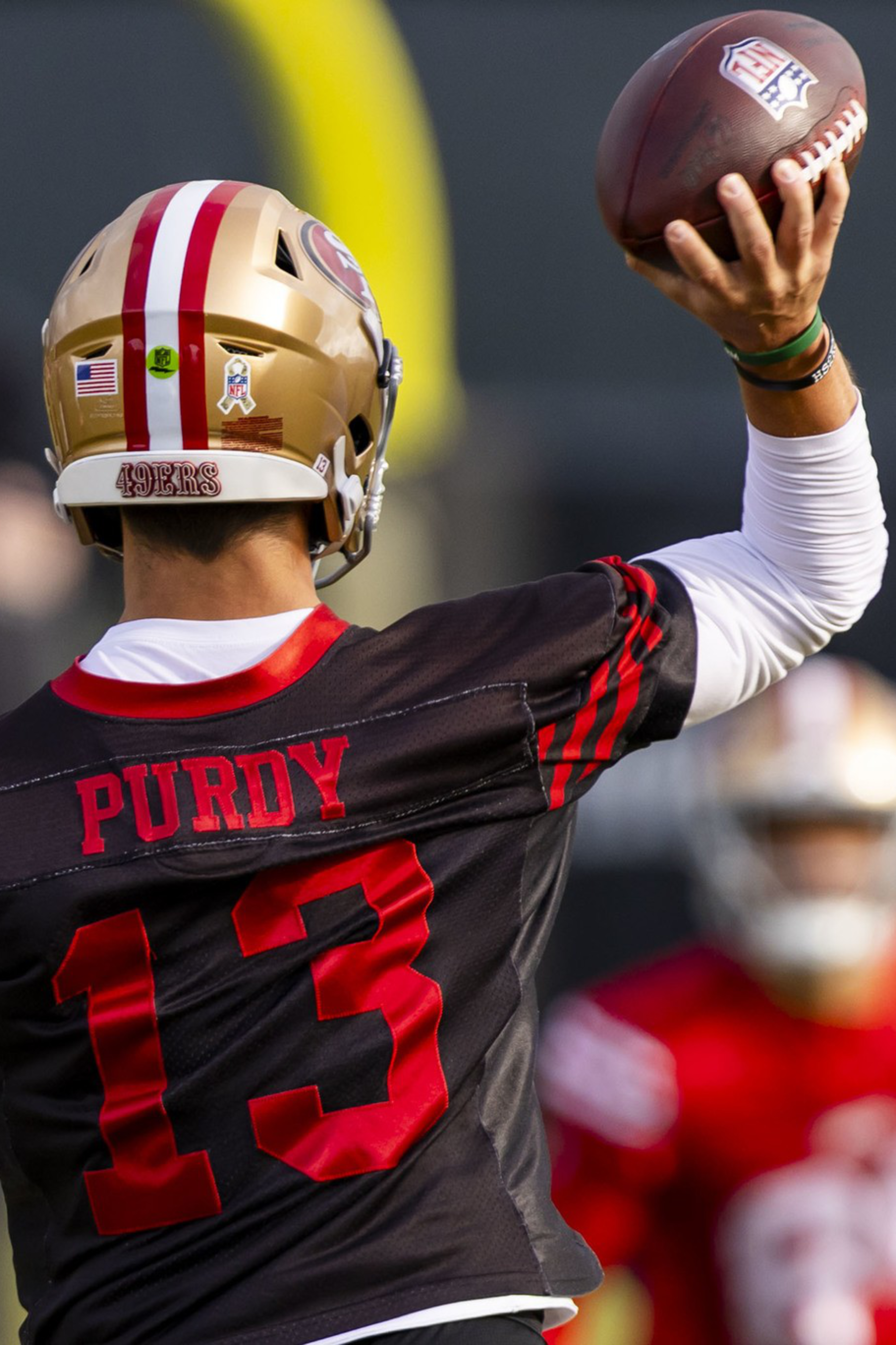 A football player wearing a black jersey with "Purdy" and number 13 prepares to throw a football during practice, with another player blurred in the background.