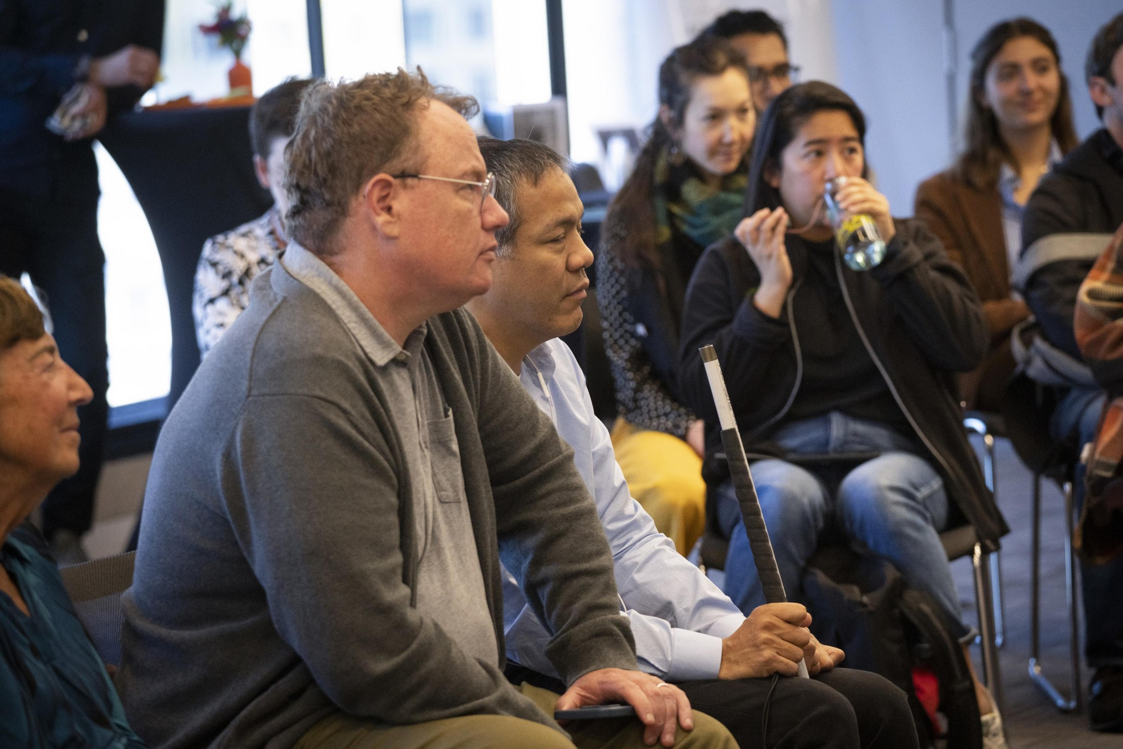 A group of people sits attentively in a room, some facing forward. One person is holding a walking cane, and another is drinking from a bottle.