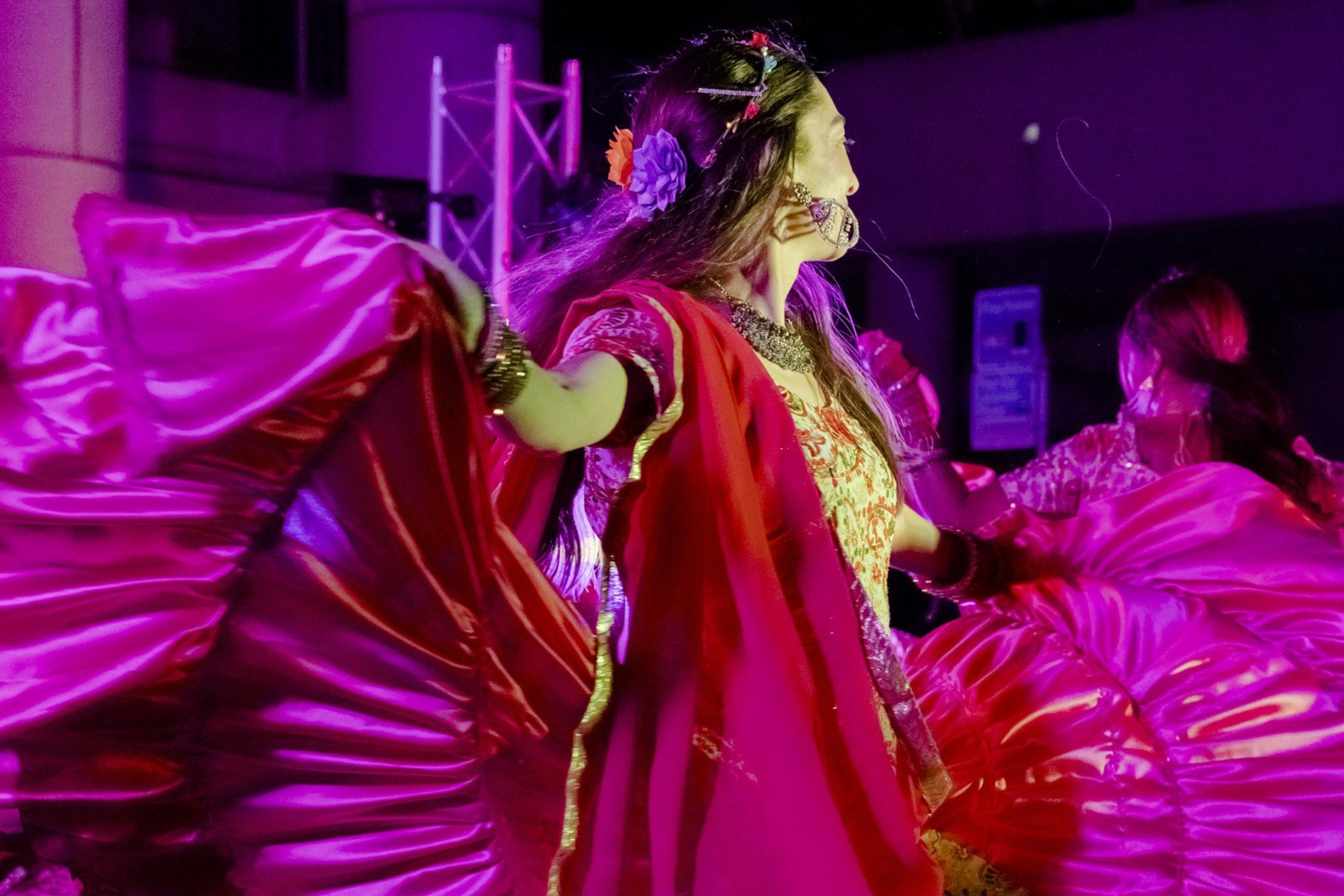 A dancer in vibrant traditional attire twirls, showcasing a wide, flowing magenta skirt under bright stage lights, creating a dynamic and colorful scene.