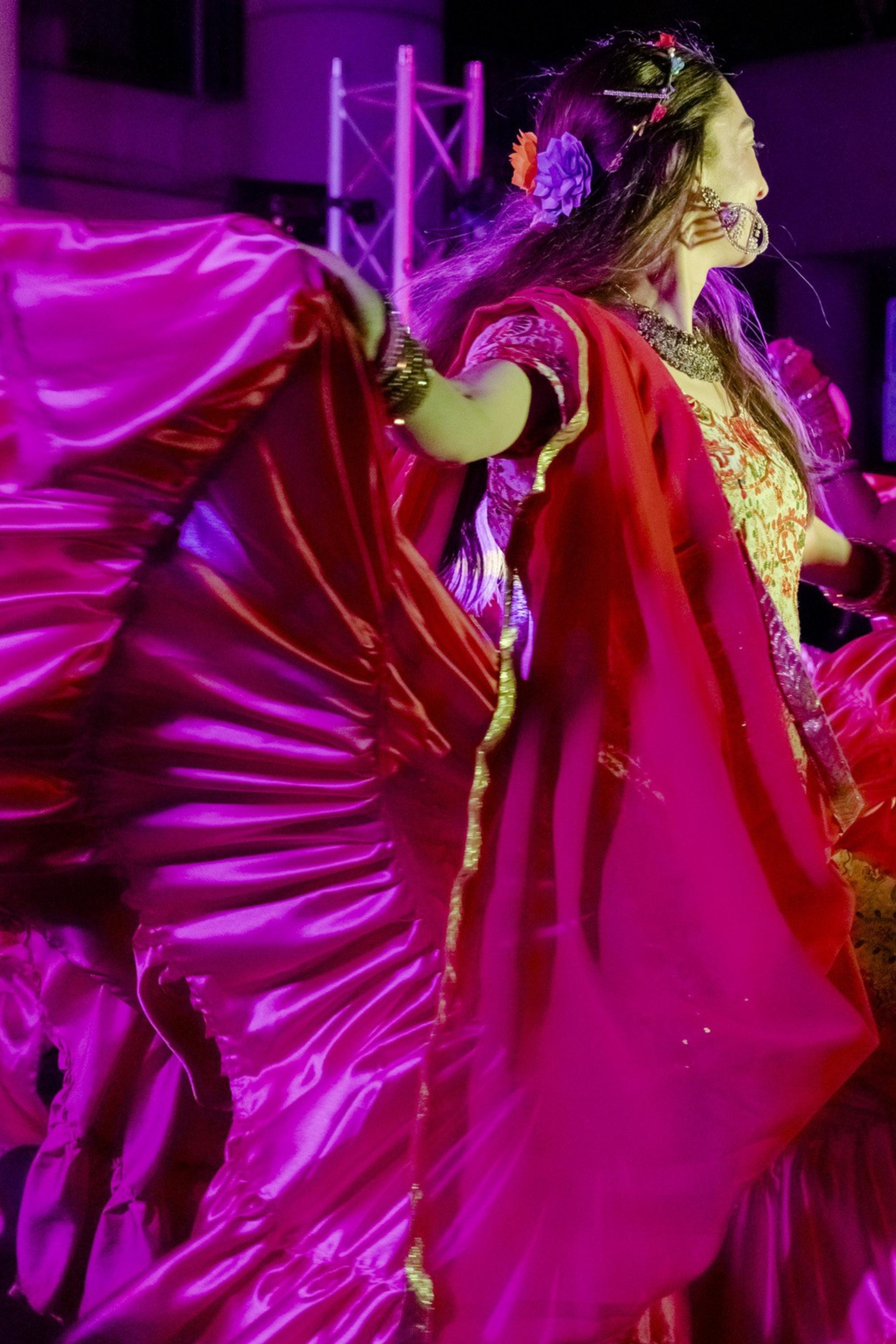 A dancer in vibrant traditional attire twirls, showcasing a wide, flowing magenta skirt under bright stage lights, creating a dynamic and colorful scene.