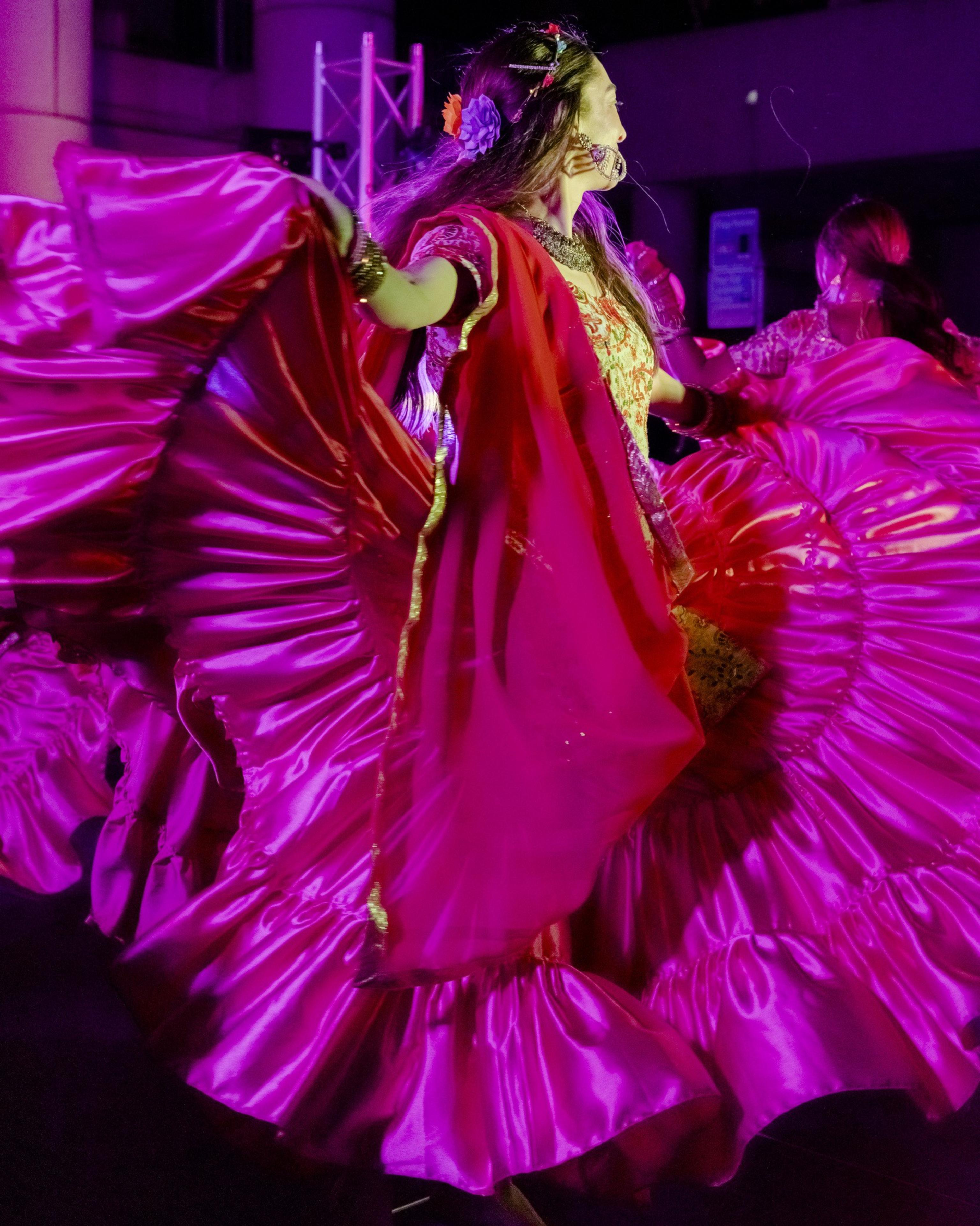 A dancer in vibrant traditional attire twirls, showcasing a wide, flowing magenta skirt under bright stage lights, creating a dynamic and colorful scene.