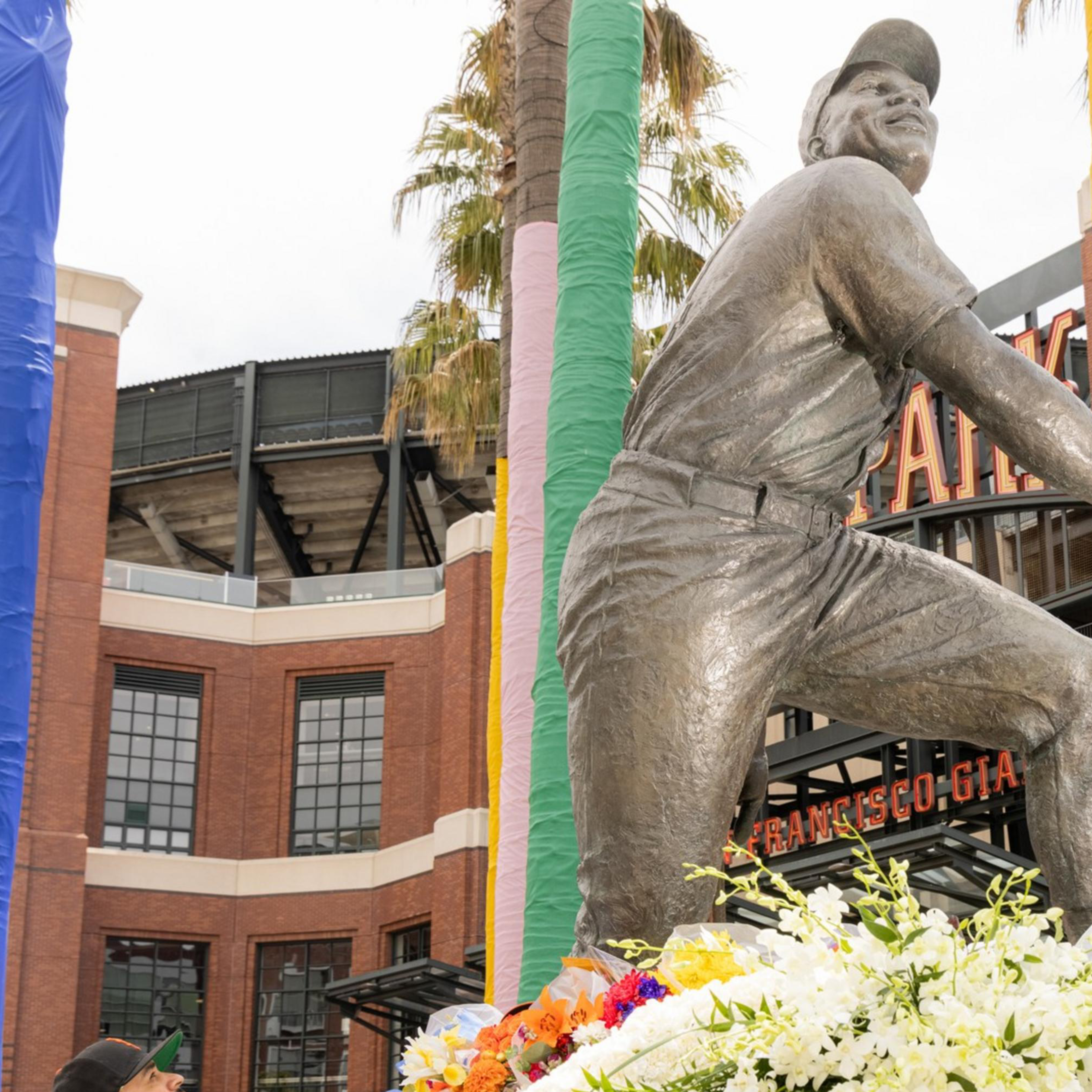 A statue of a baseball player is surrounded by colorful wrapped poles and flowers. A man wearing a baseball jersey is adjusting a floral display at the statue's base.