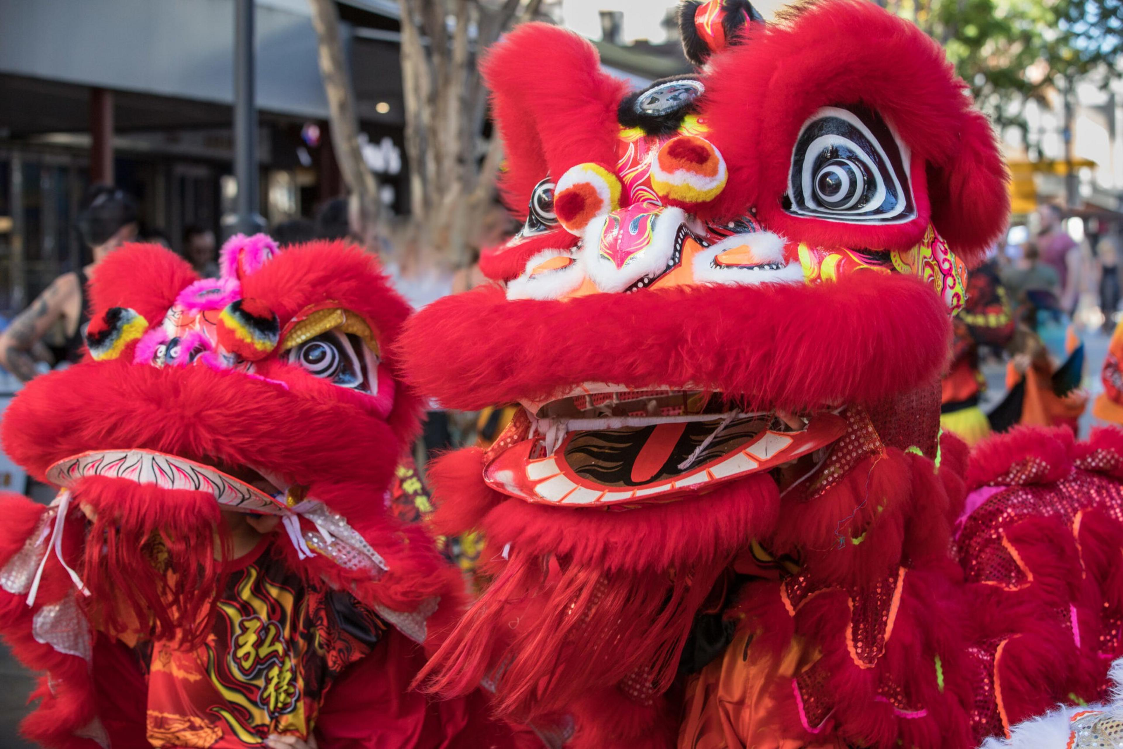 Two vibrant red lion dance costumes with colorful details are worn by performers during a lively outdoor festival.