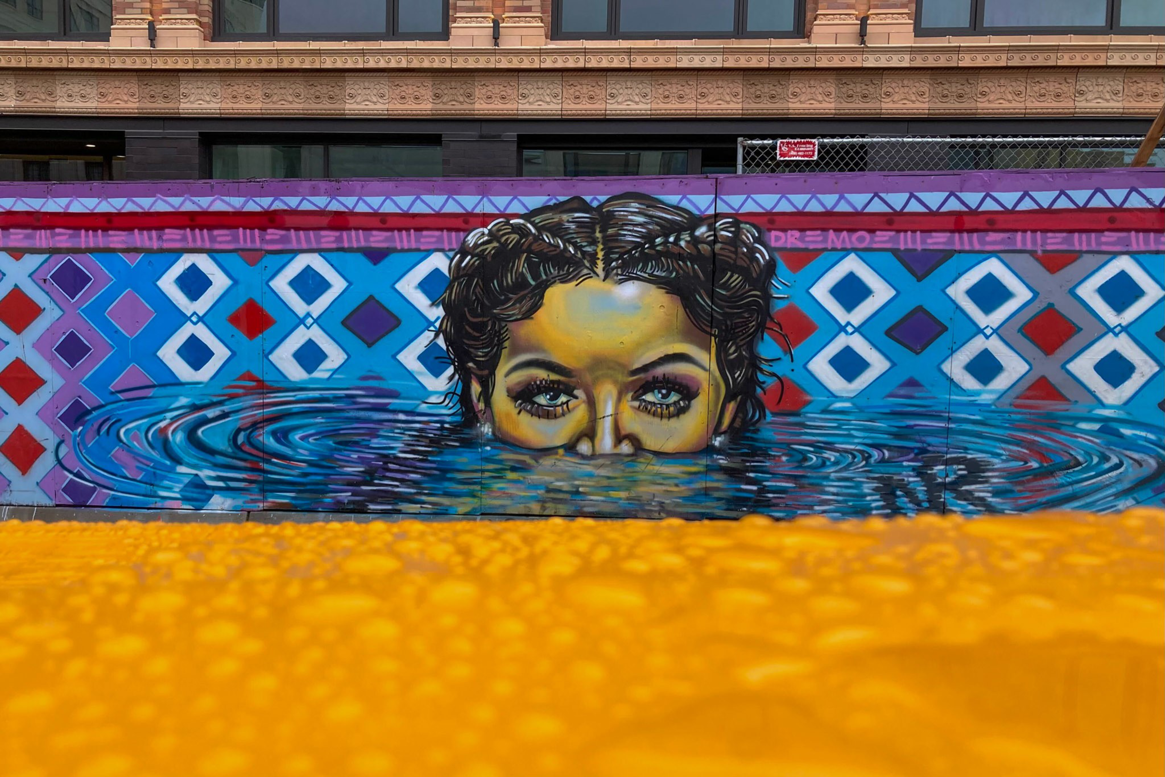 A mural depicts a woman's face with braided hair emerging from water, set against a blue patterned background with geometric shapes.