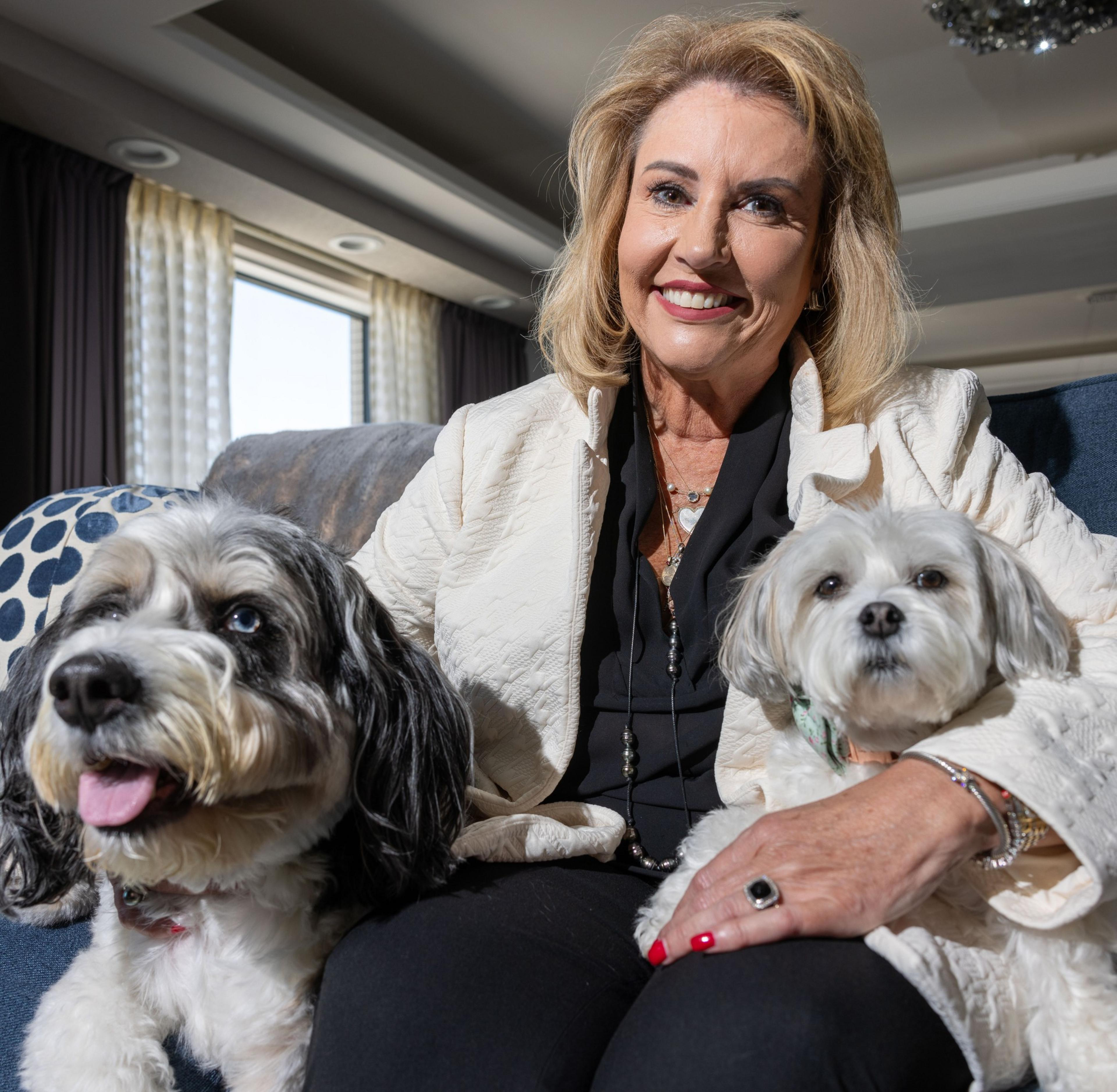 A smiling woman with blond hair, wearing a cream-colored jacket and black top, sits indoors on a couch with two small dogs, one black and white, the other white.