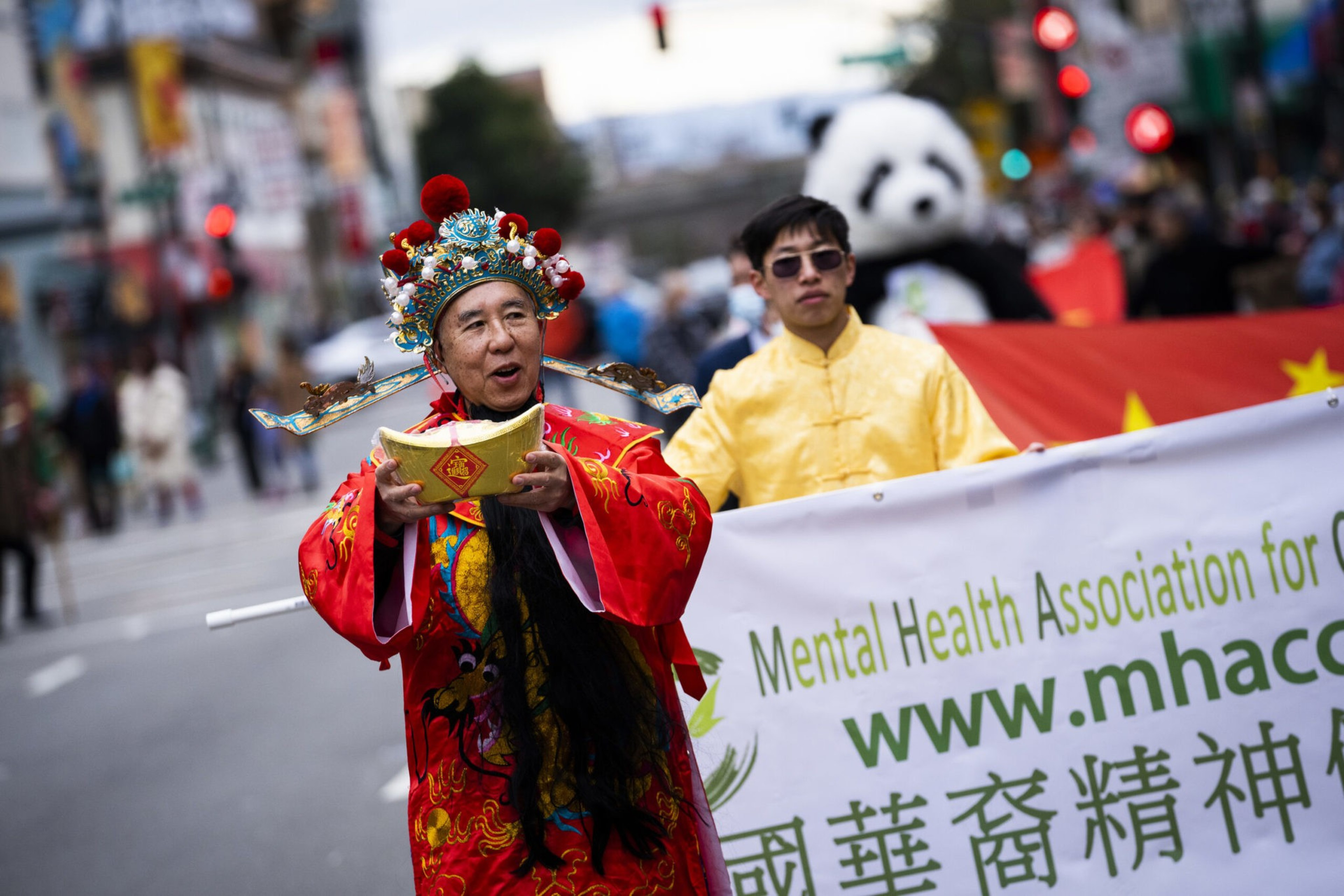 A man in a colorful traditional Chinese costume holds a gold ingot, followed by another man in yellow and a banner from the Mental Health Association.