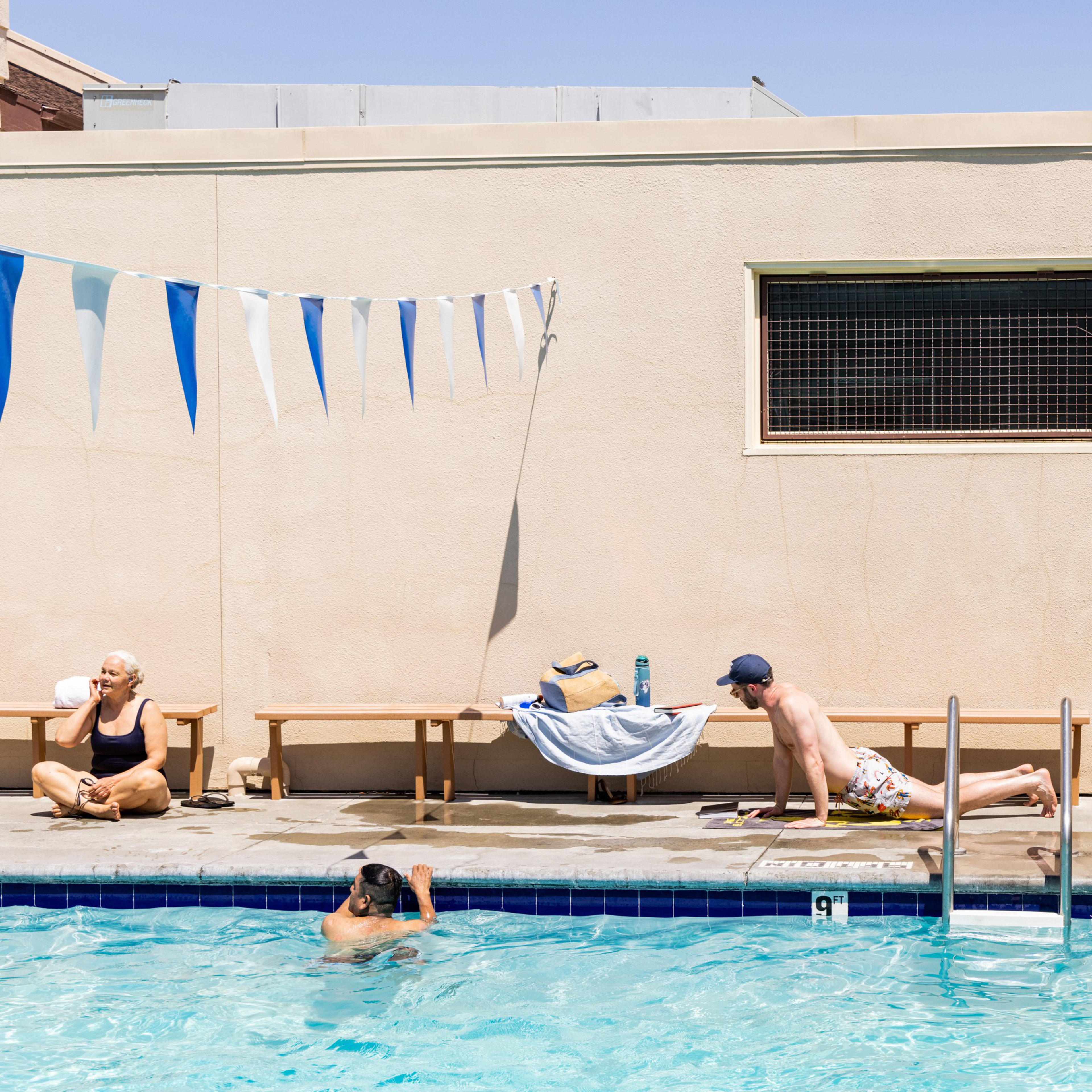Three people are at a pool; one is swimming, one is doing push-ups, and one is sitting on the deck near a wall with blue and white flag bunting above.