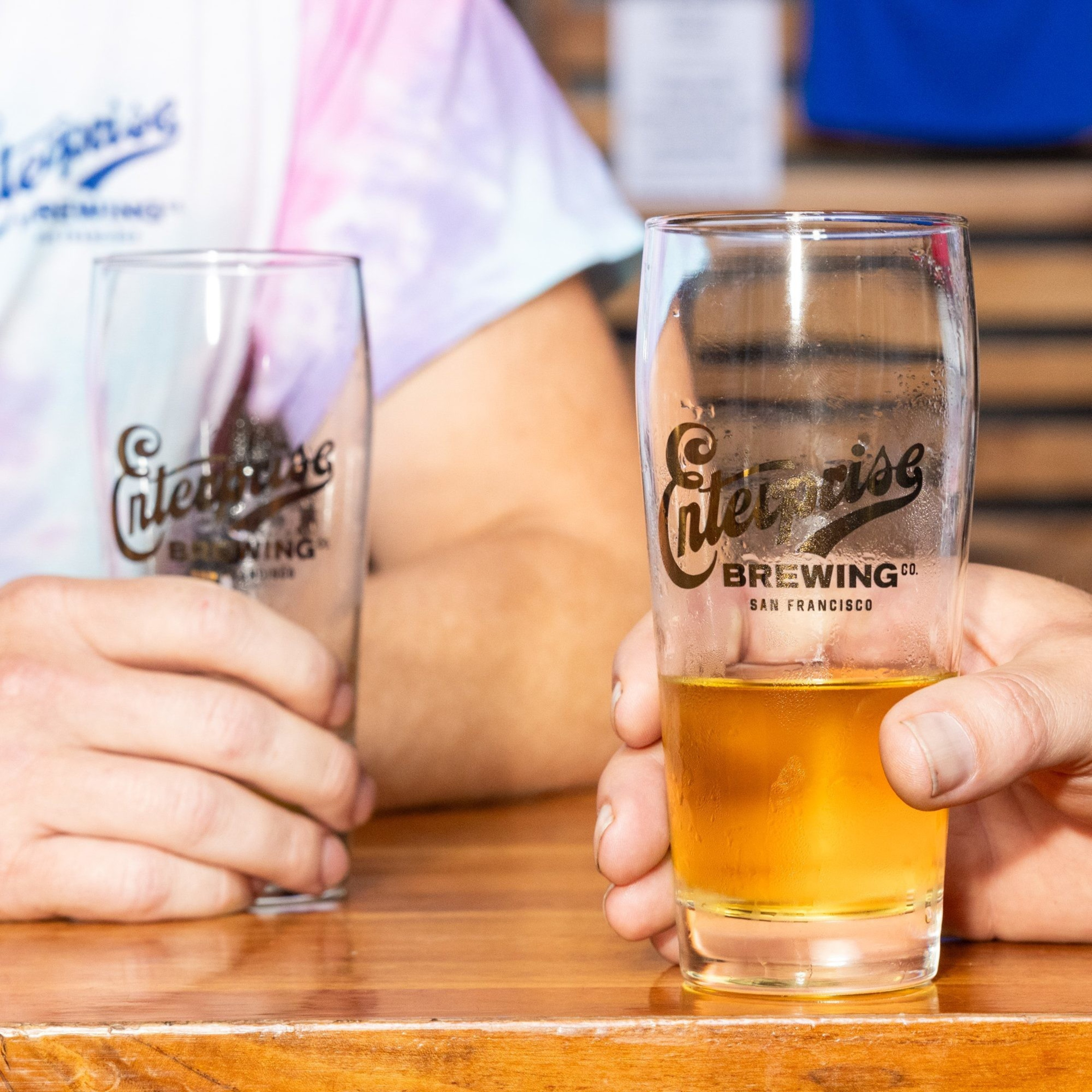 Two people hold glasses of beer with the &quot;Enterprise Brewing Co. San Francisco&quot; logo, seated at a wooden table. One glass is half full, the other nearly empty.