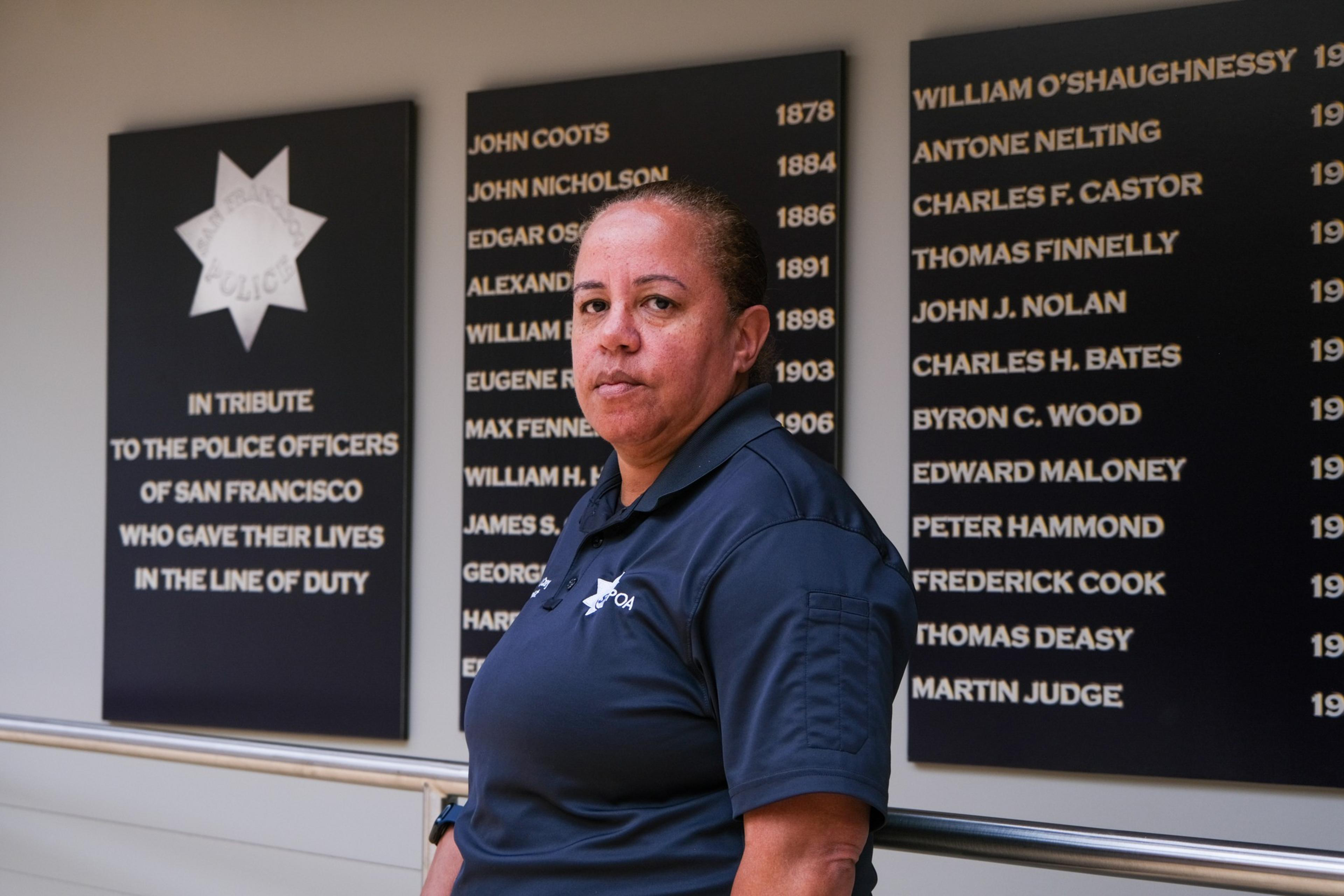A woman in a police uniform stands solemnly before a memorial wall honoring fallen San Francisco officers.