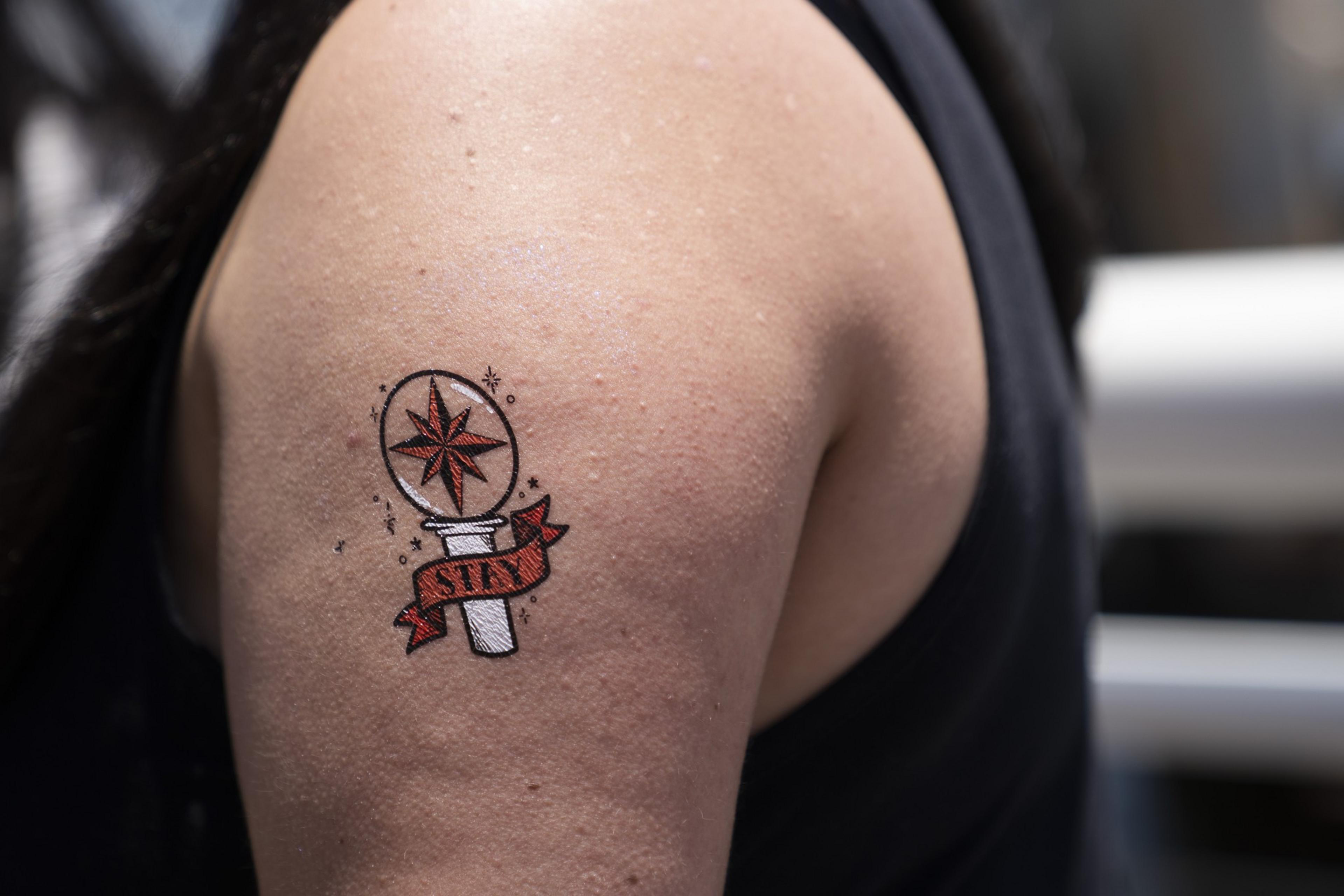 The image shows an arm with a tattoo of a red star and compass inside a circle, a ribbon with &quot;STAY&quot; wrapped around the base, surrounded by small stars.