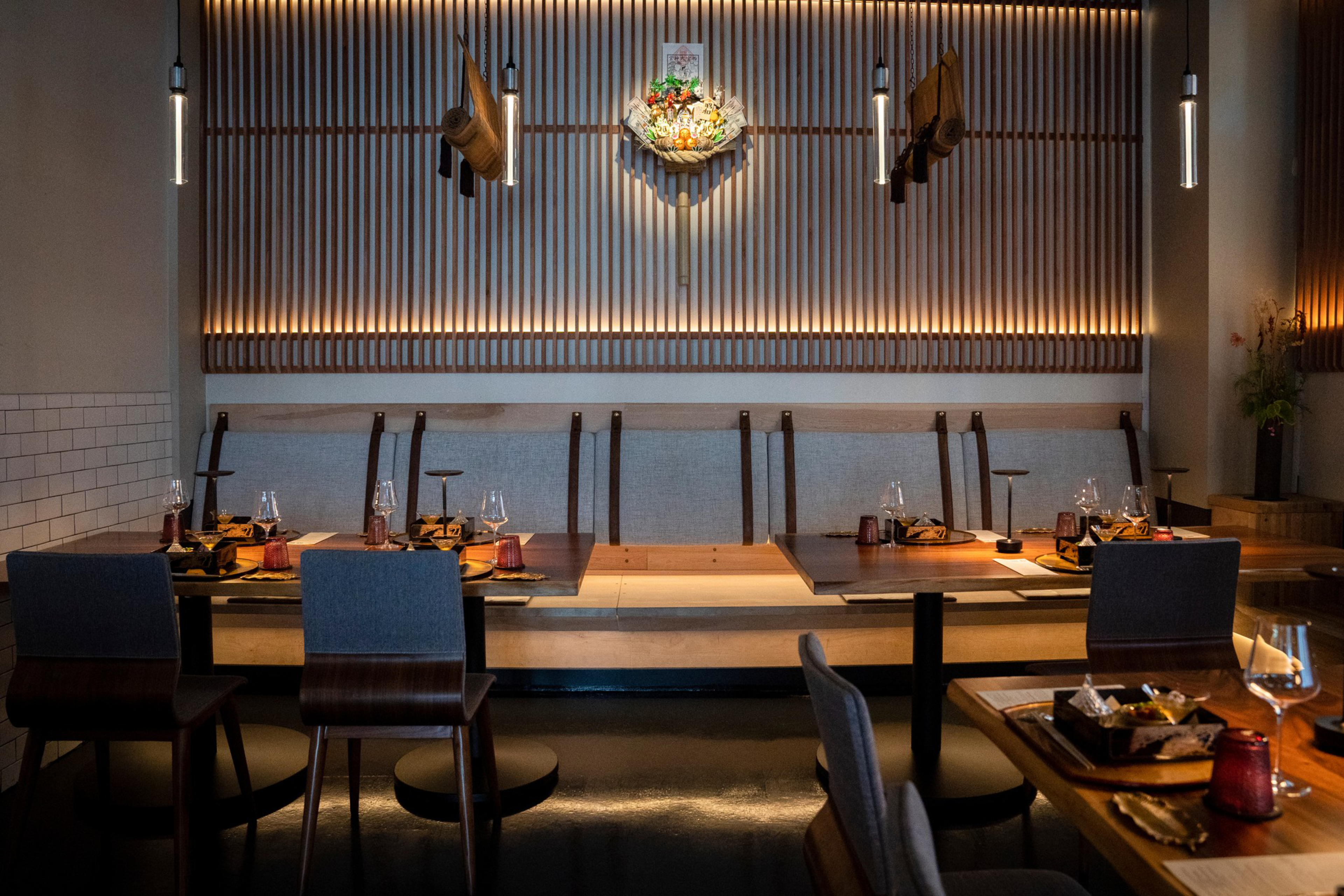 A modern dining area with wooden tables, blue chairs, and a backlit slatted wood wall decor featuring hanging items. Each table is set with glassware and plates.